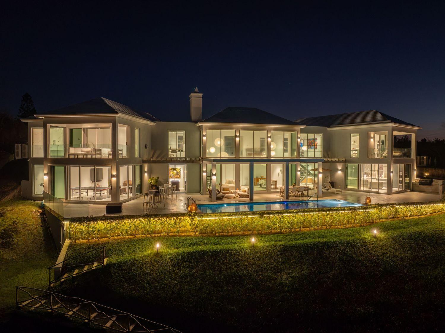 Modern luxury home exterior illuminated at night with warm lighting and landscaped gardens. Drone photogrpahy.