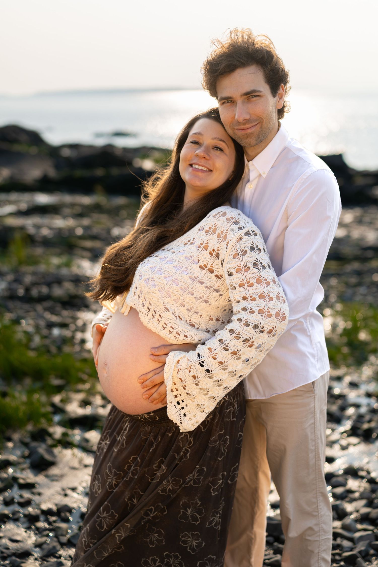Couple embracing on rocky beach at sunset, pregnant woman in lace top cradling baby bump with partner in white shirt.