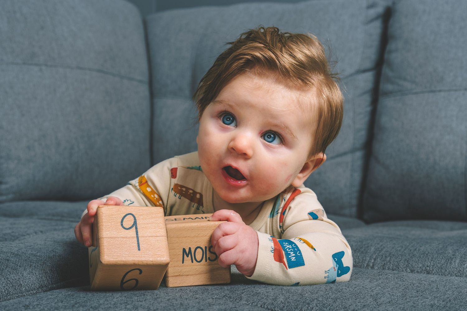 A baby in a patterned onesie plays with wooden alphabet blocks on a gray couch.