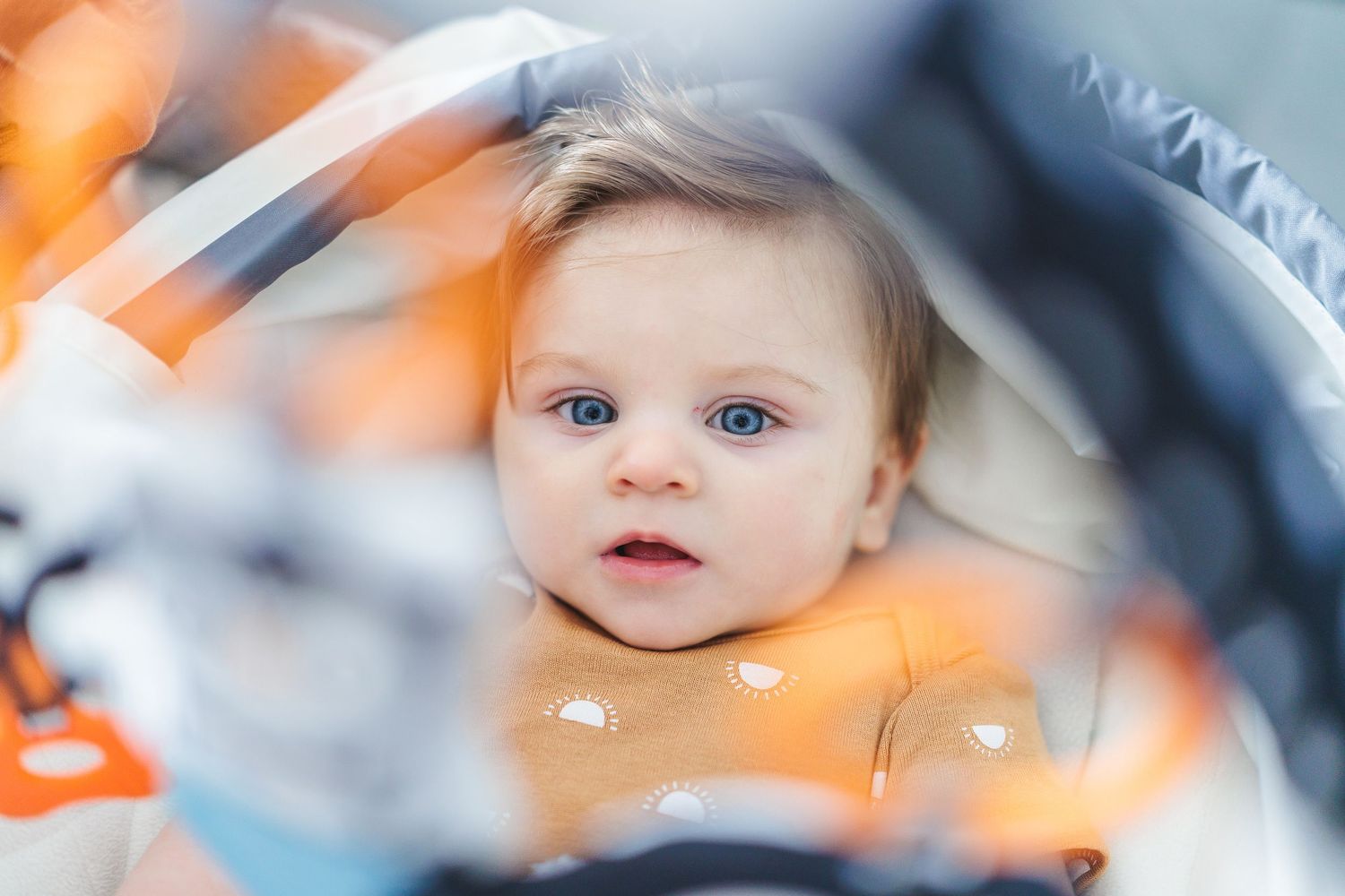 A baby with bright blue eyes gazes through blurred orange foreground elements.