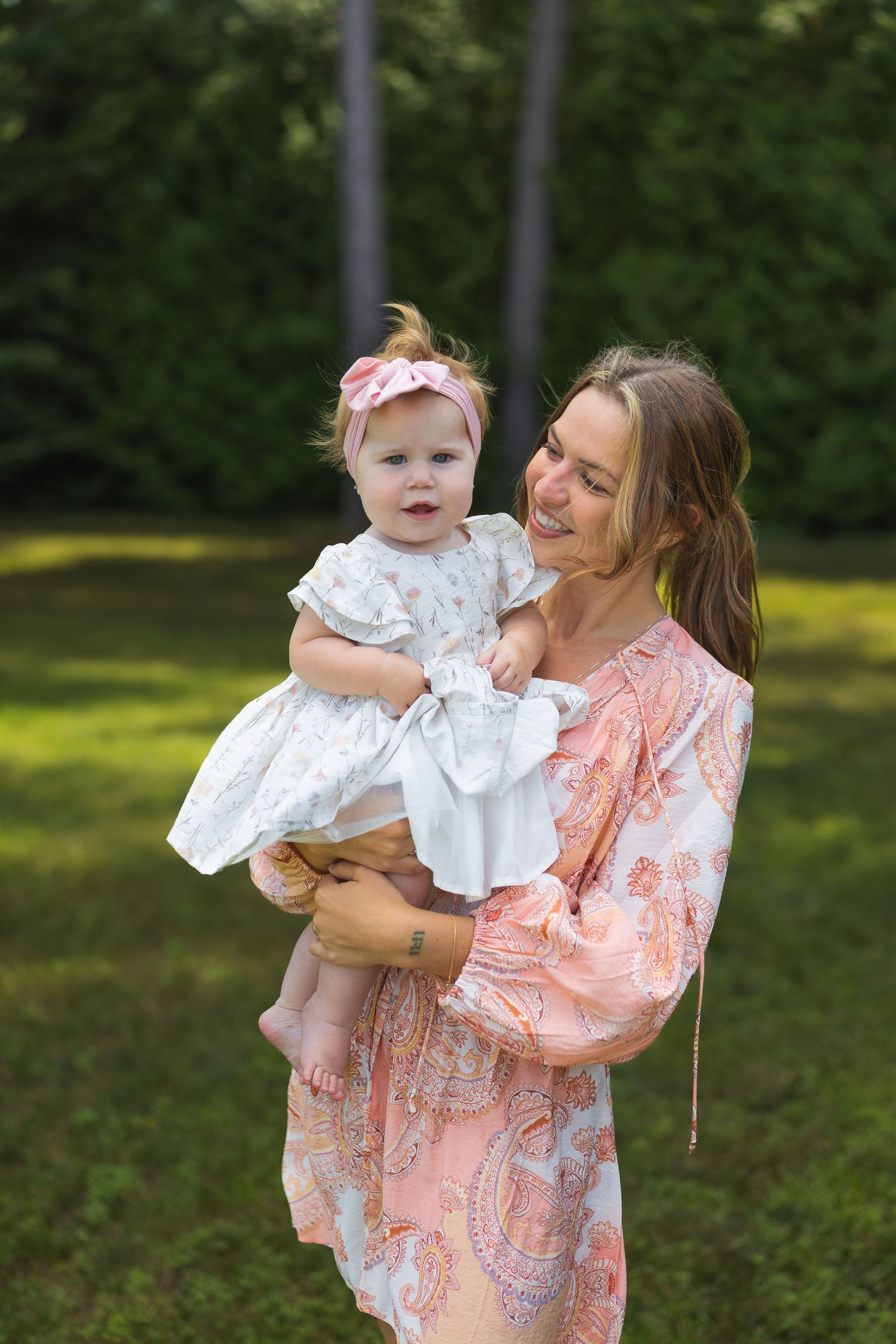 A smiling woman in a pink paisley dress holds a baby girl in a white dress with a pink bow headband outdoors.