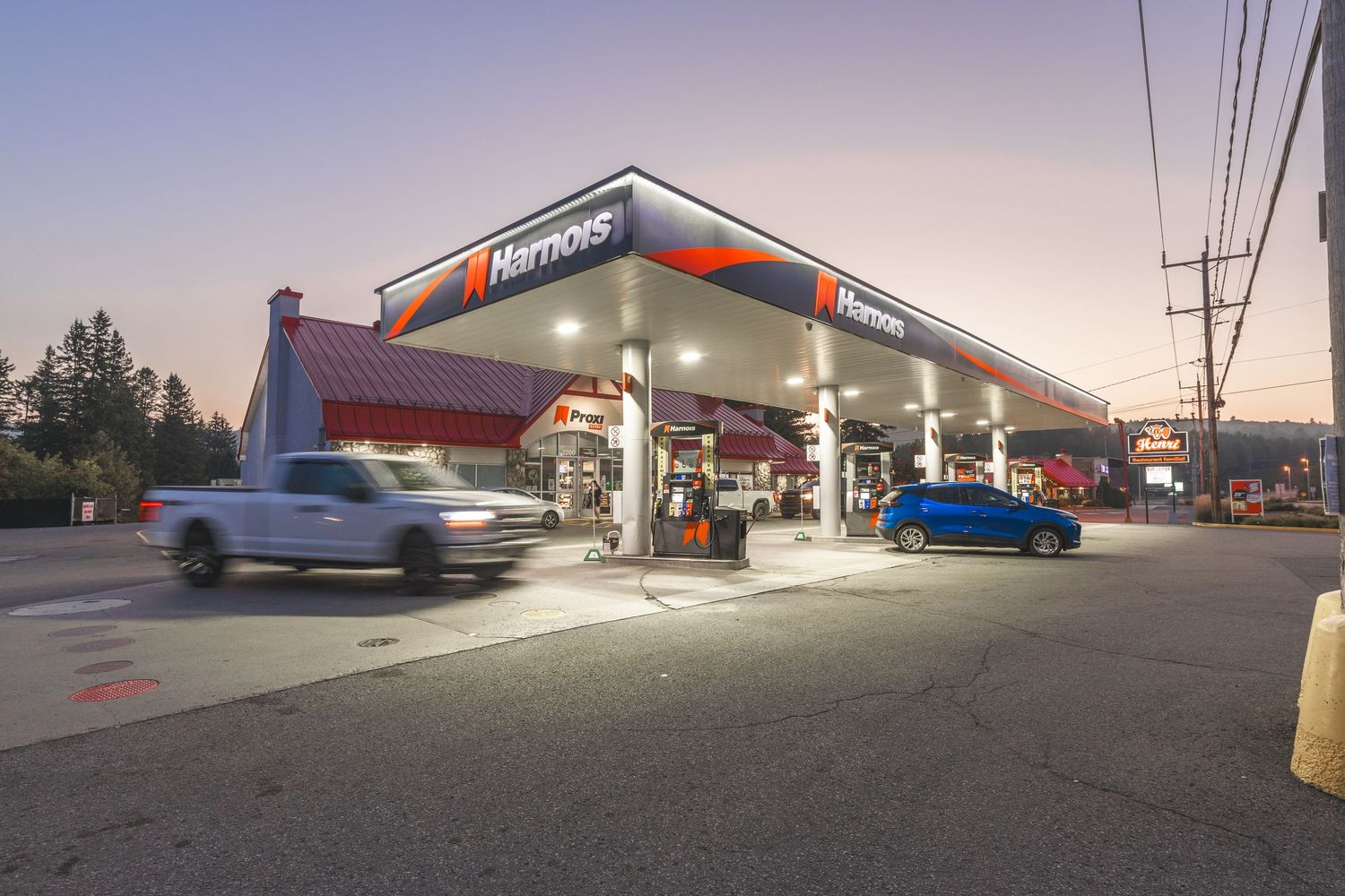 A panoramic view of a gas station convenience store at dusk with illuminated signage and parked vehicles.