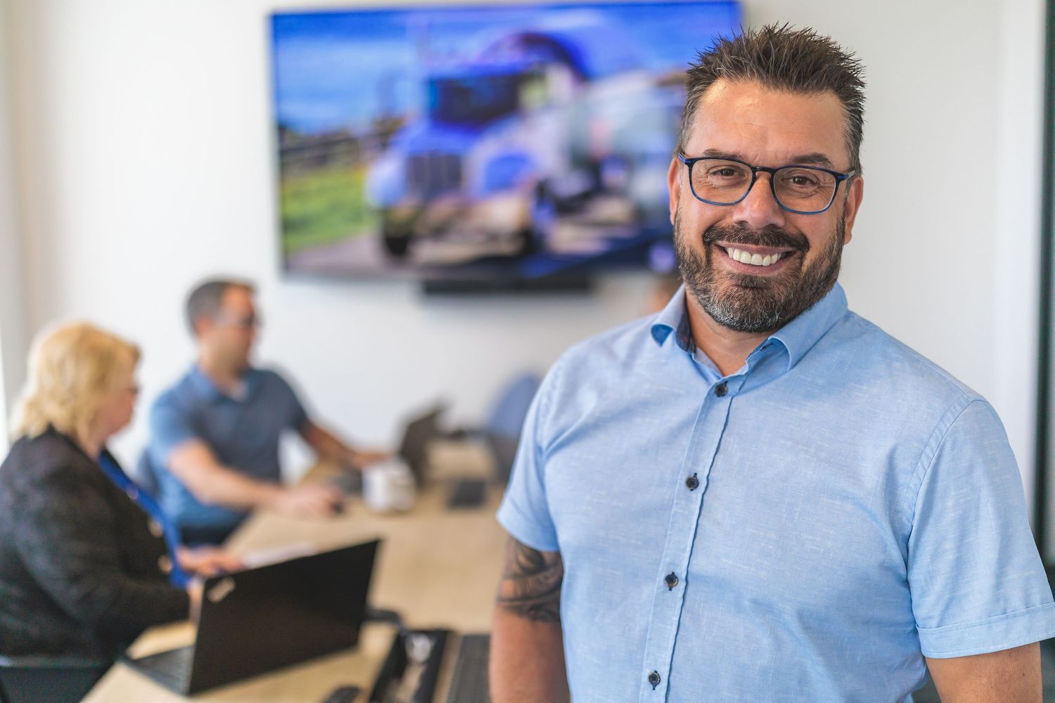 Professional in light blue shirt smiles in office with meeting room visible in background.