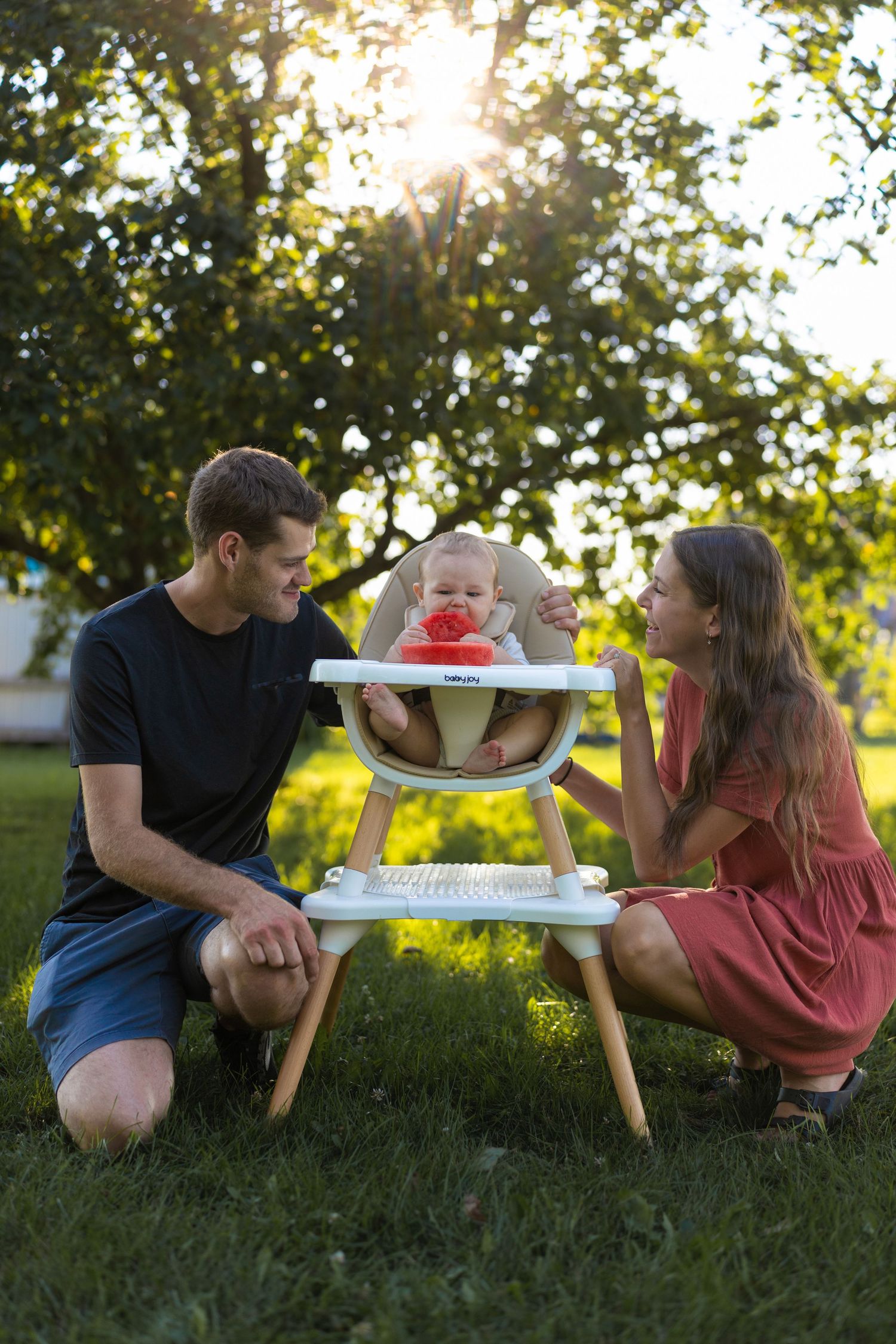 Parents feeding baby in high chair outdoors in sunny garden during golden hour family moment.