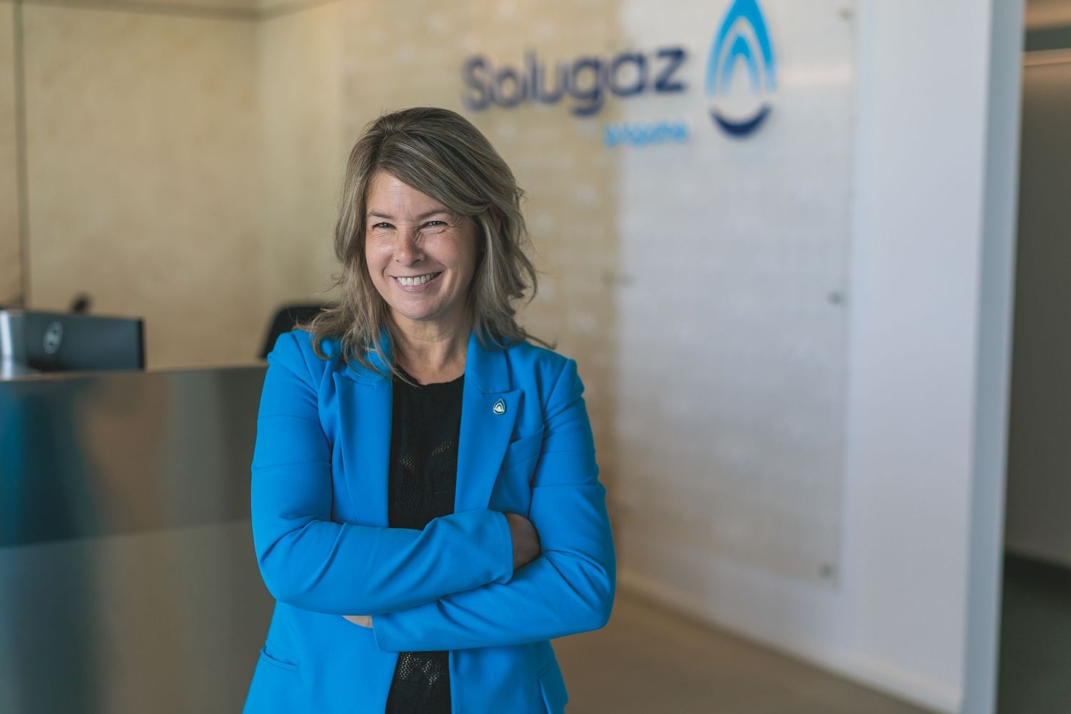 Professional in bright blue blazer stands against office wall with company logo.