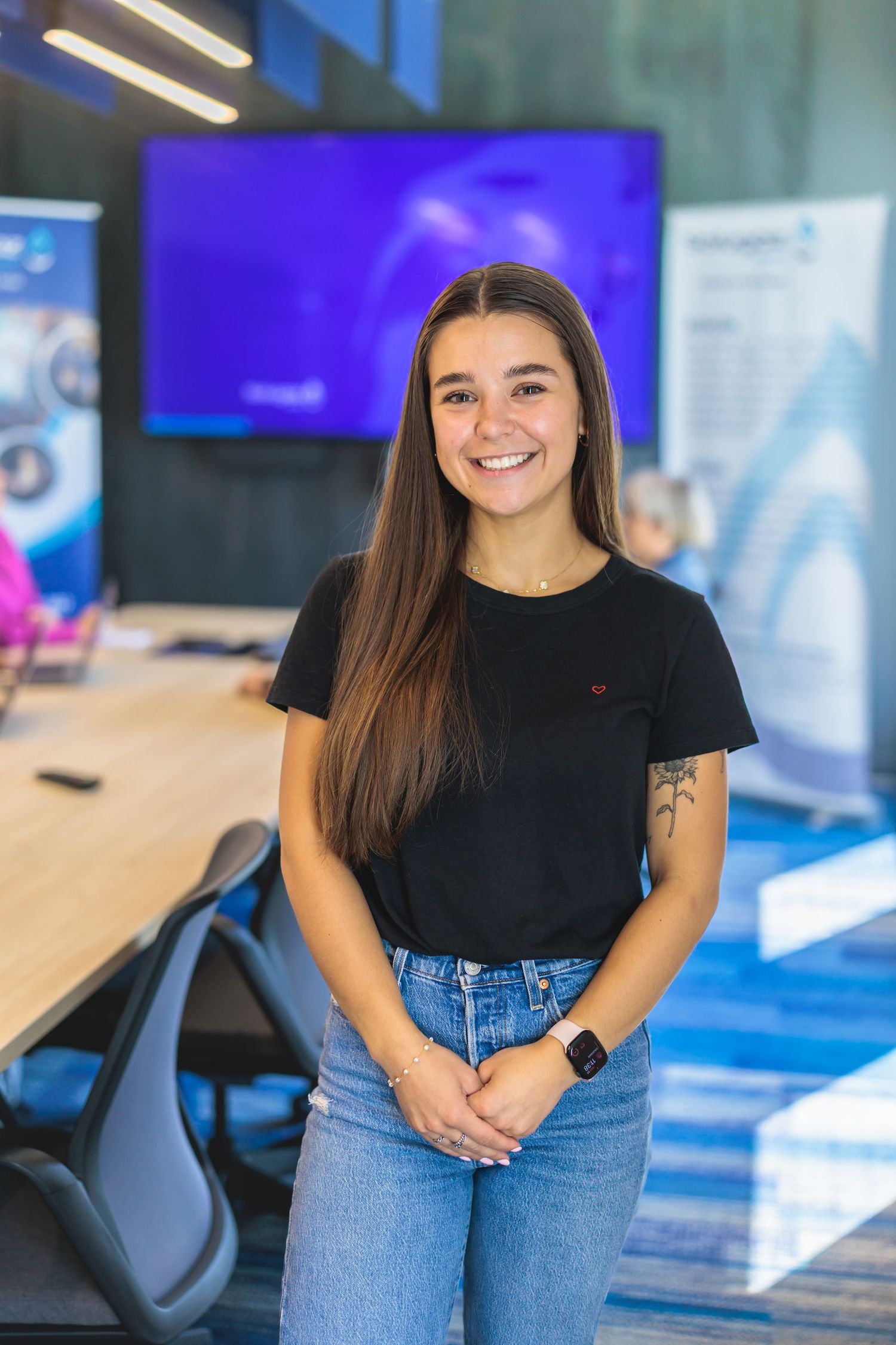 Professional in casual attire poses in modern office space with blue display screen.