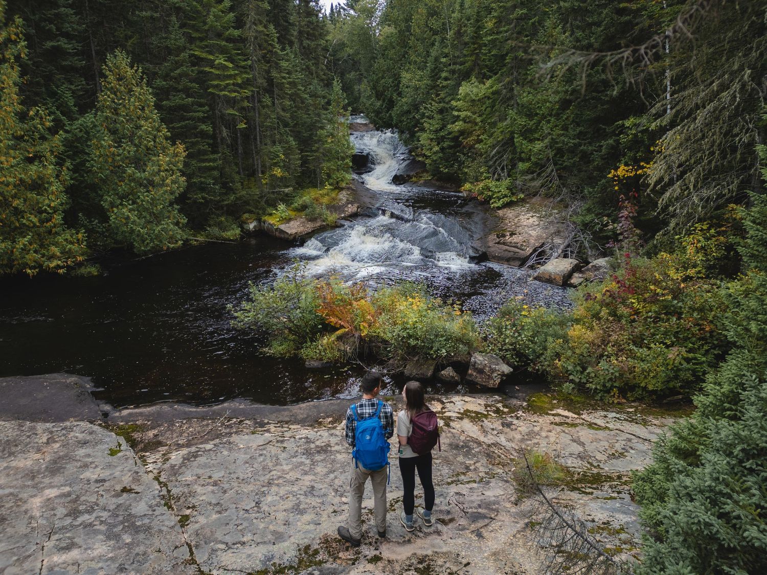 People hiking along rocky riverbank surrounded by lush forest landscape.