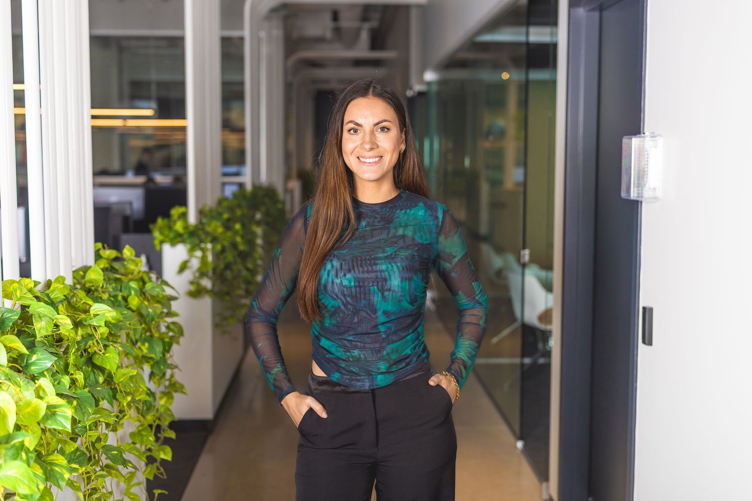 Professional headshot in office hallway with indoor plants and natural lighting.