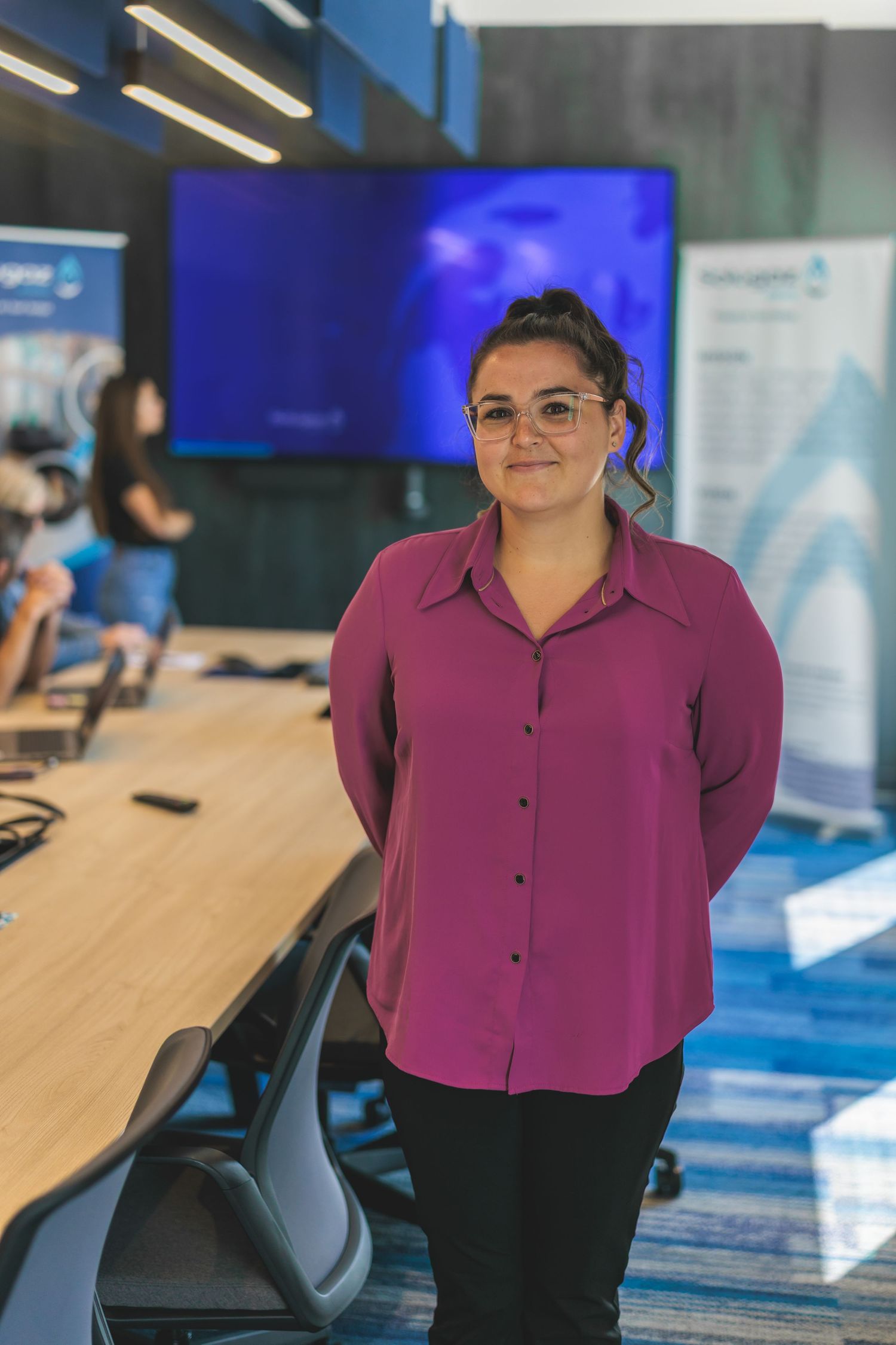 Employee in magenta blouse stands in modern office meeting room with blue display screen.