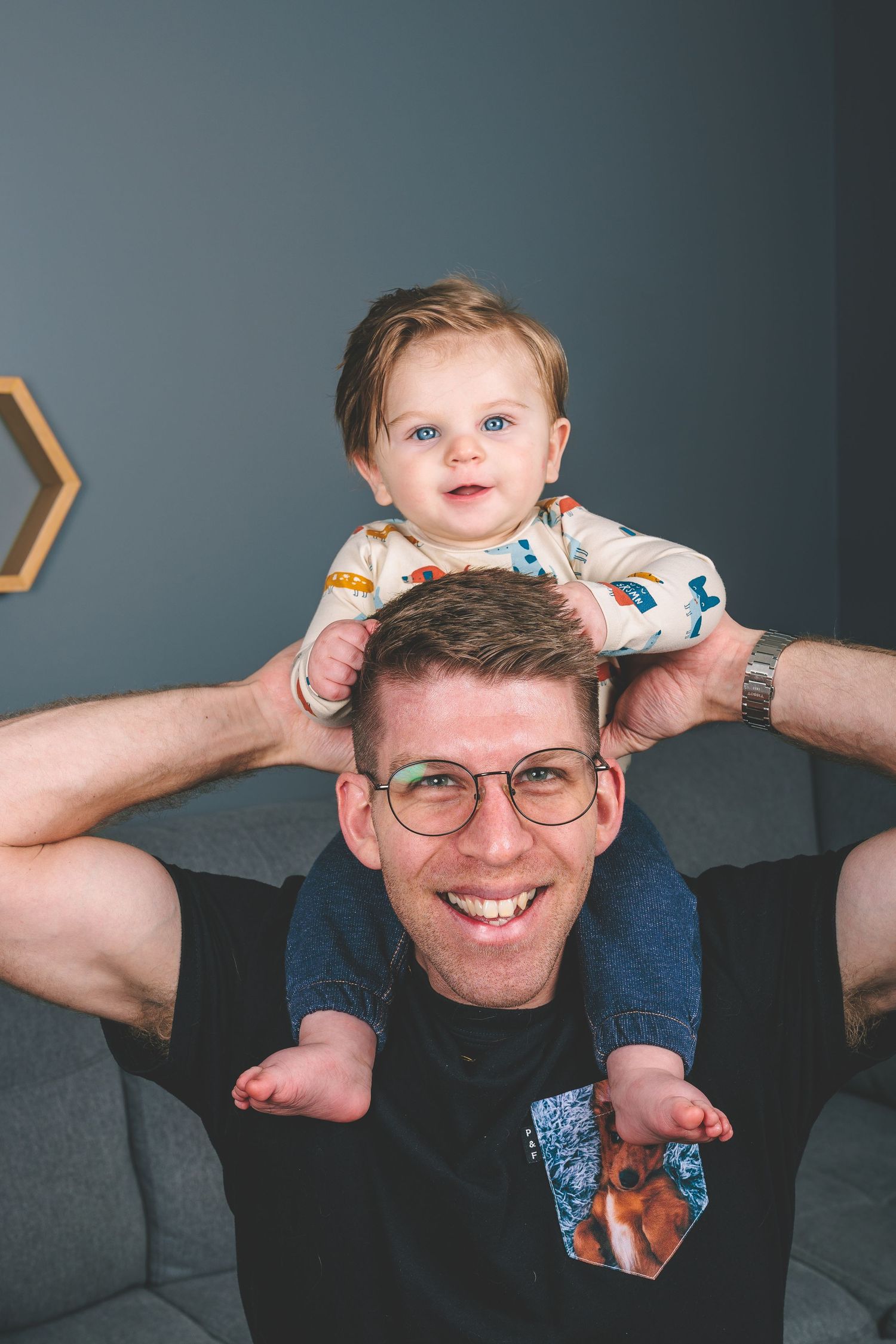 A joyful shoulder ride moment between parent and child against a gray wall with hexagon shelves.