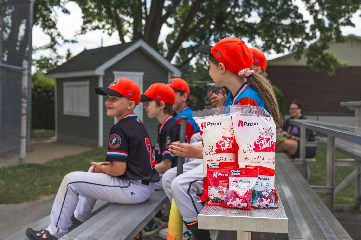 Baseball players in red caps sit in a dugout with stacks of Budweiser beer boxes visible behind them.