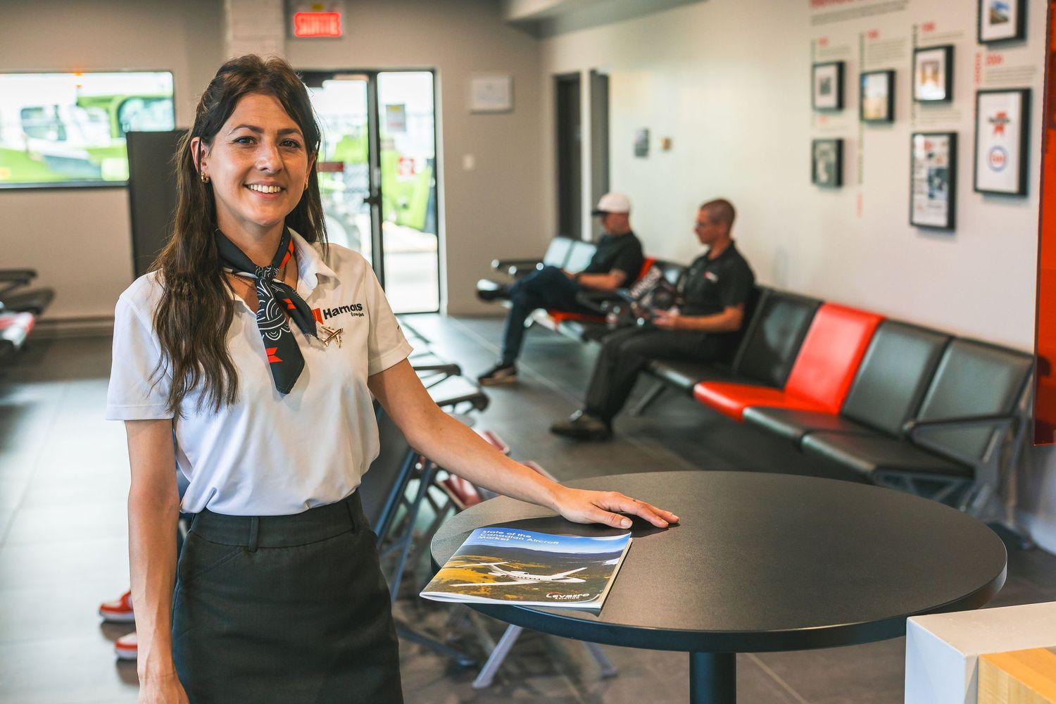 Professional in a white polo shirt stands at reception desk while others wait in office lobby.
