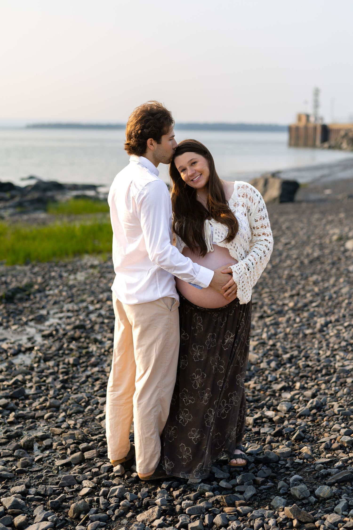 A pregnant couple embraces on a rocky beach at sunset, the woman smiling warmly while her partner holds her baby bump.