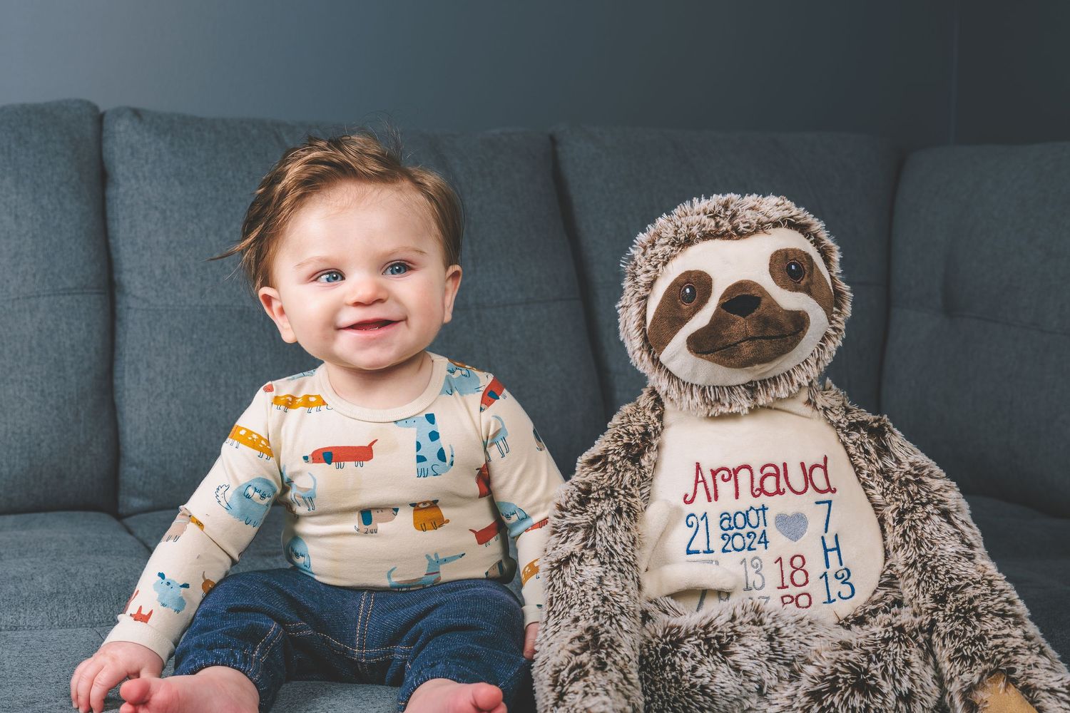 A smiling toddler sits on a gray couch next to a plush sloth stuffed animal.