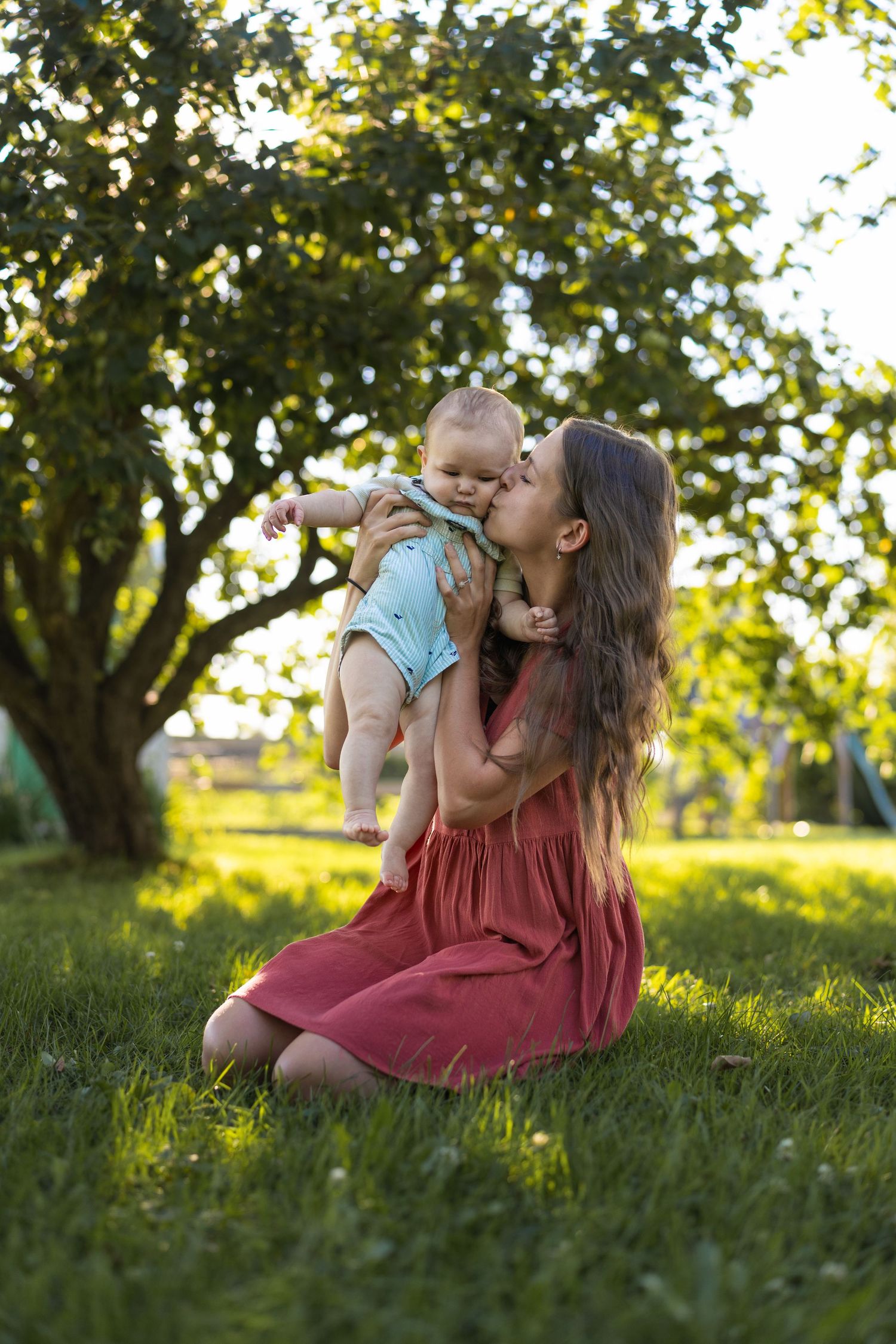 A young mother in a red dress sits on green grass, lovingly kissing her baby while holding them up outdoors.