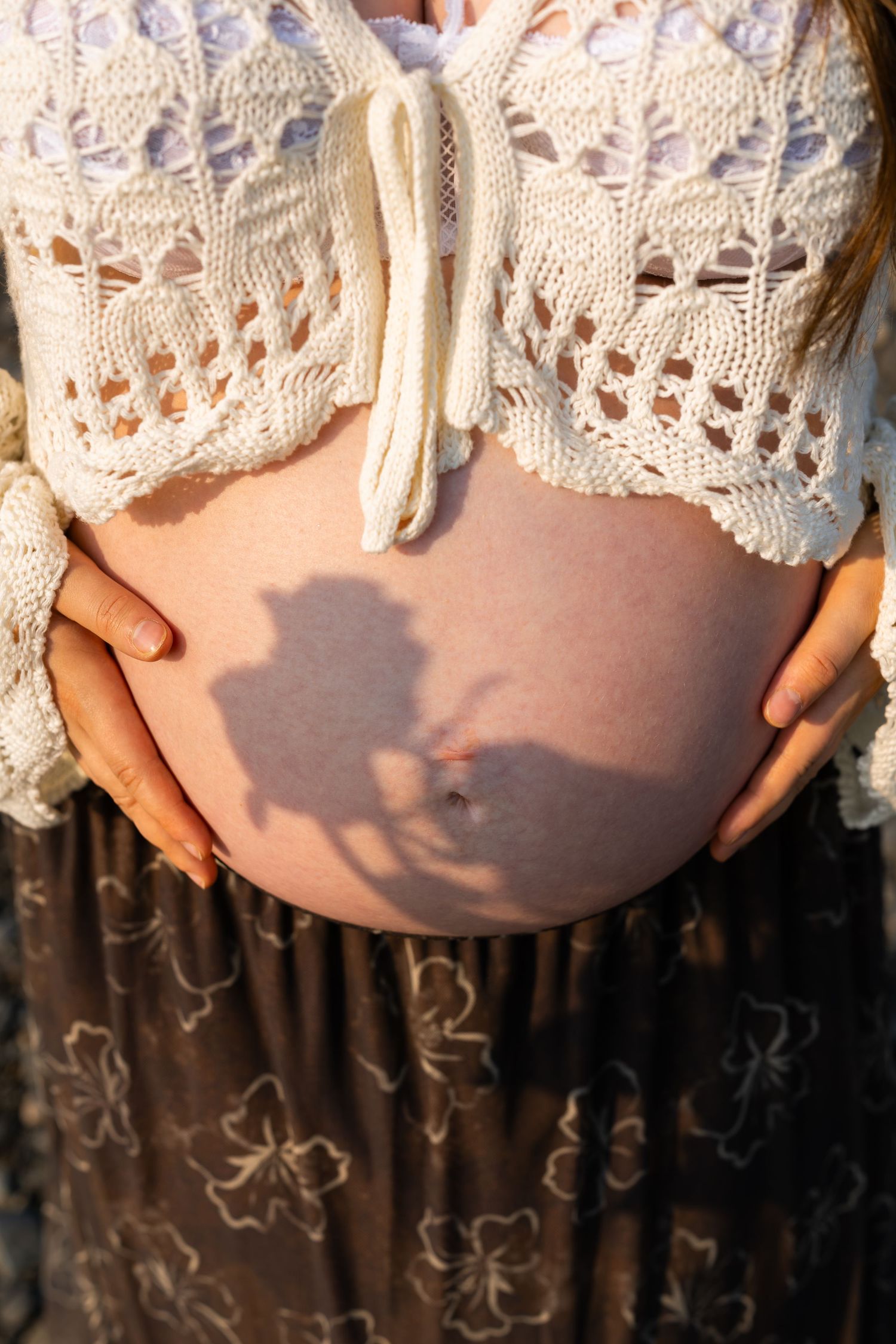 Close-up of a pregnant woman's bare belly, hands cradling bump, wearing a white knit crop top and floral skirt outdoors.