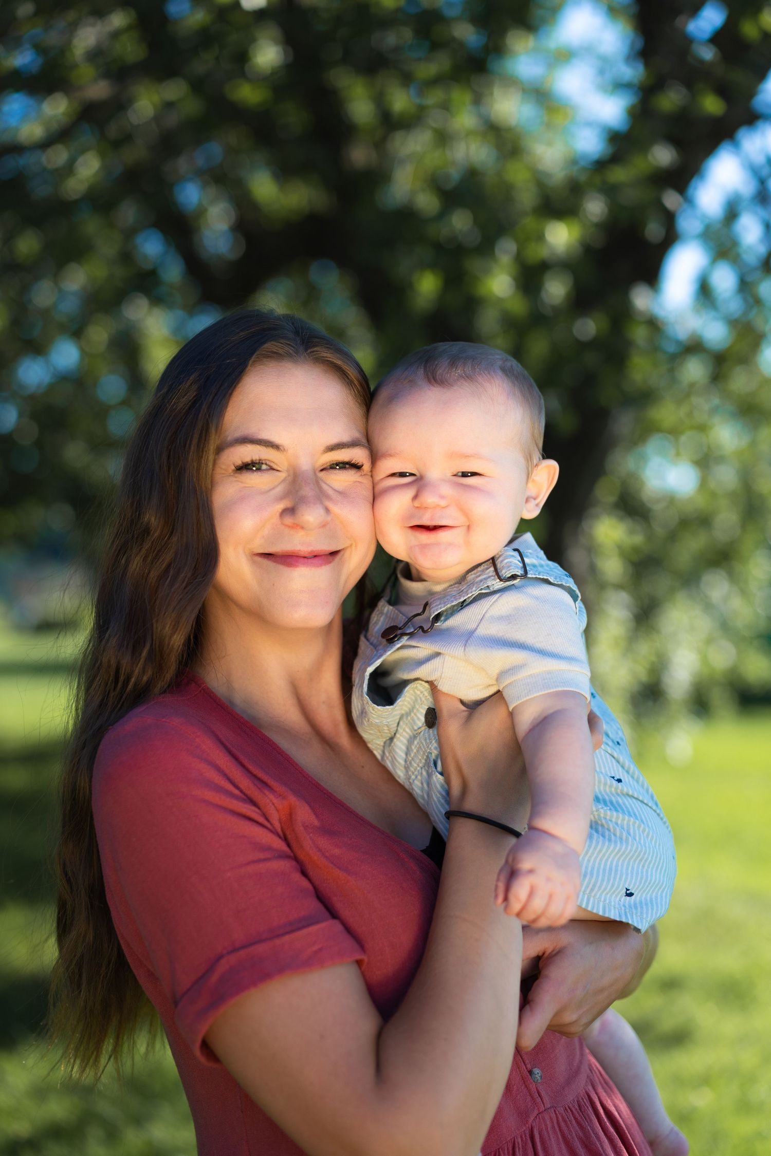Young woman in red dress smiling while holding a happy baby boy outdoors in a sunny green park.
