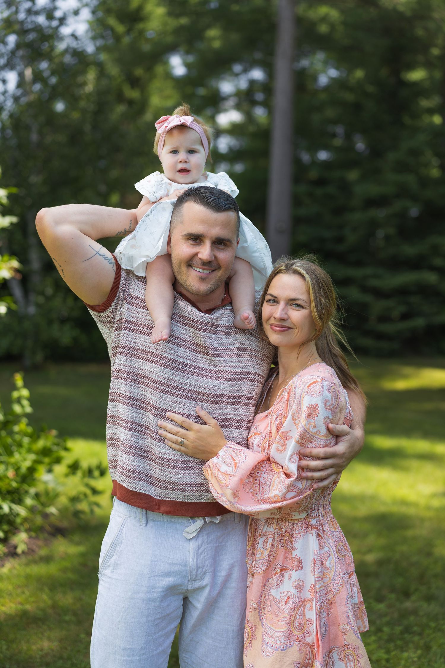Happy family portrait outdoors, man with baby on shoulders hugging smiling woman in pink dress, green trees background.