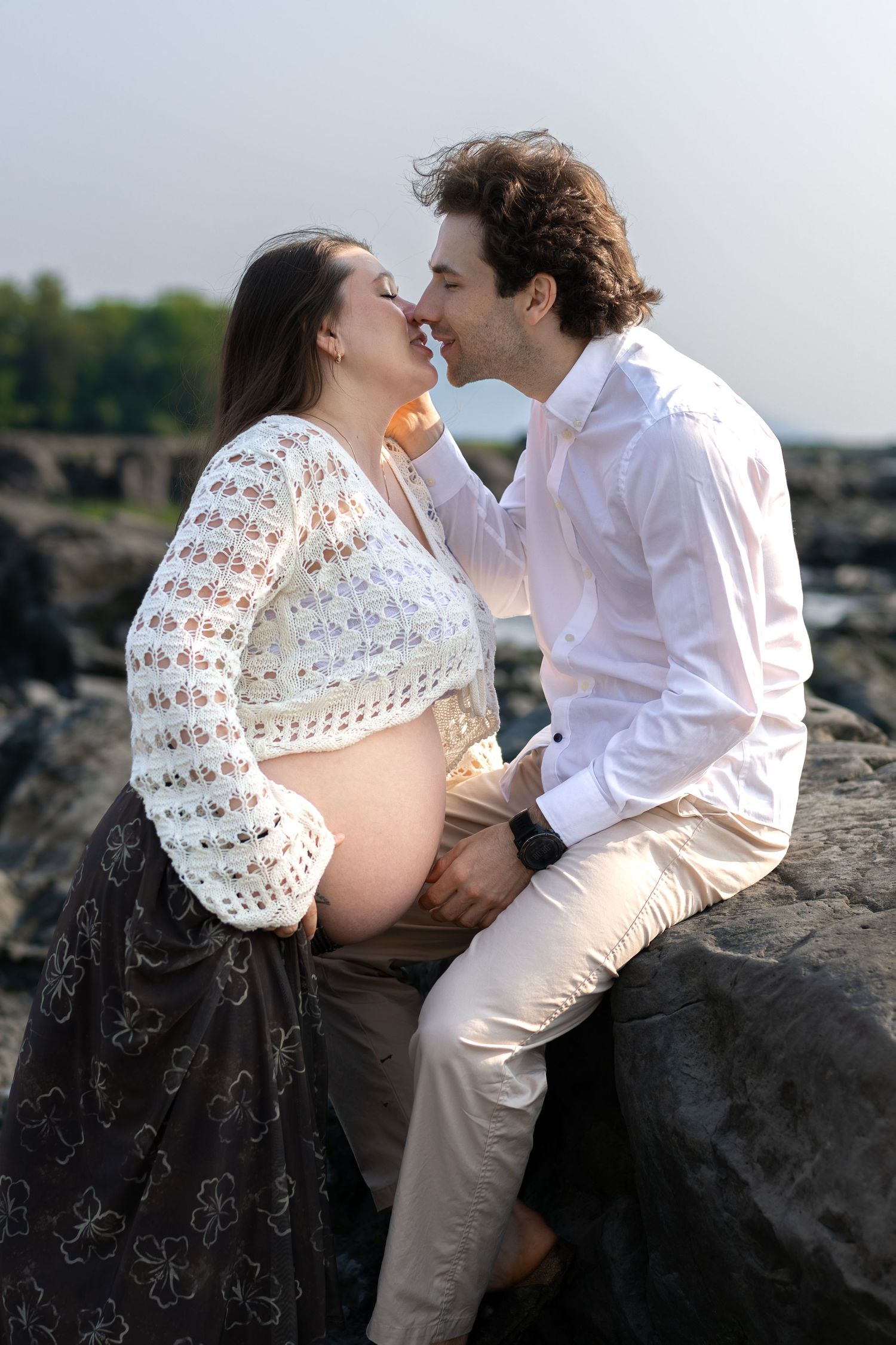 Pregnant couple sharing a tender moment on rocky shore, man in white shirt holding baby bump while leaning in for a kiss.