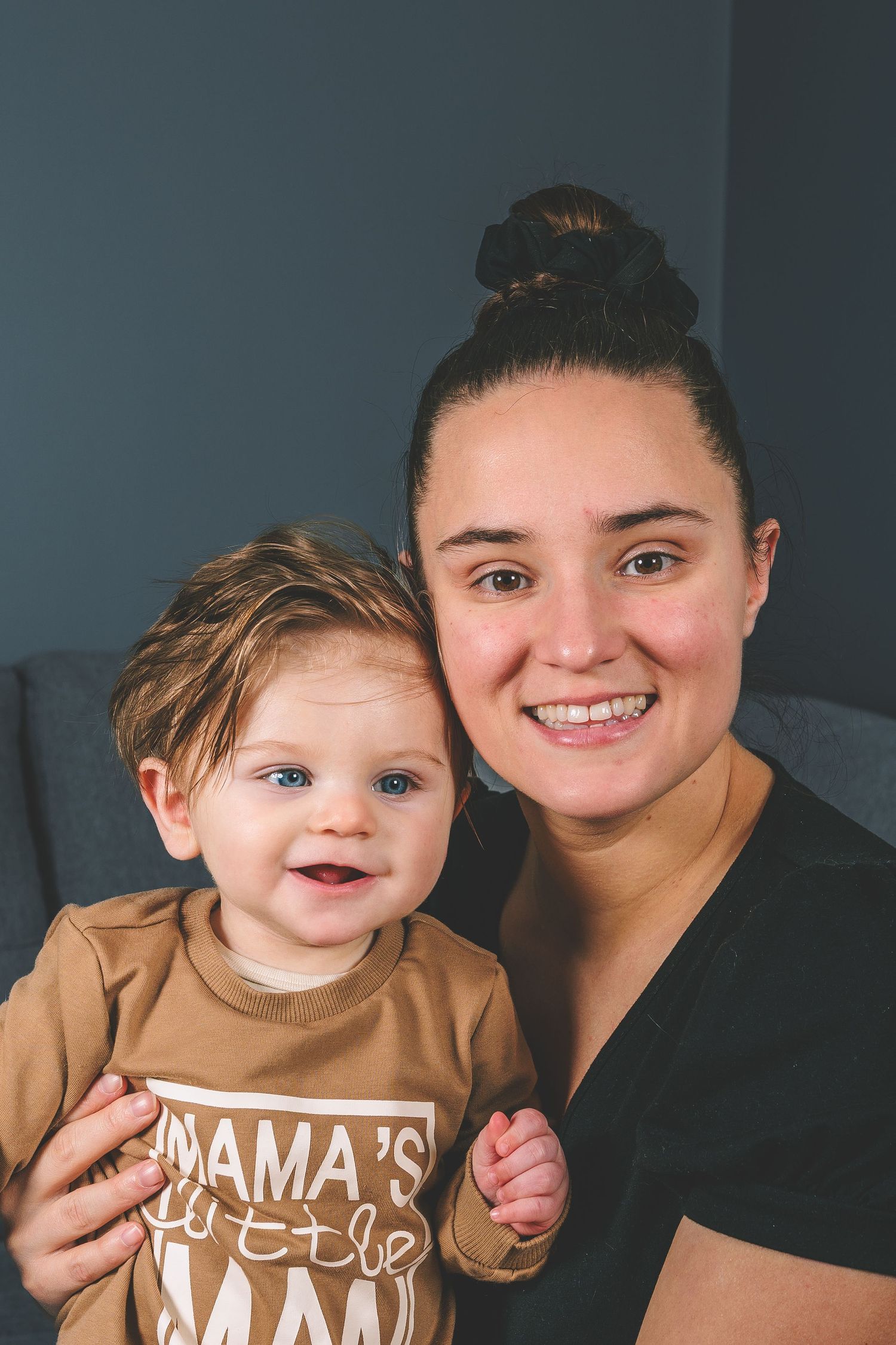 A joyful parent and baby in matching outfits share a heartwarming moment on a gray couch.