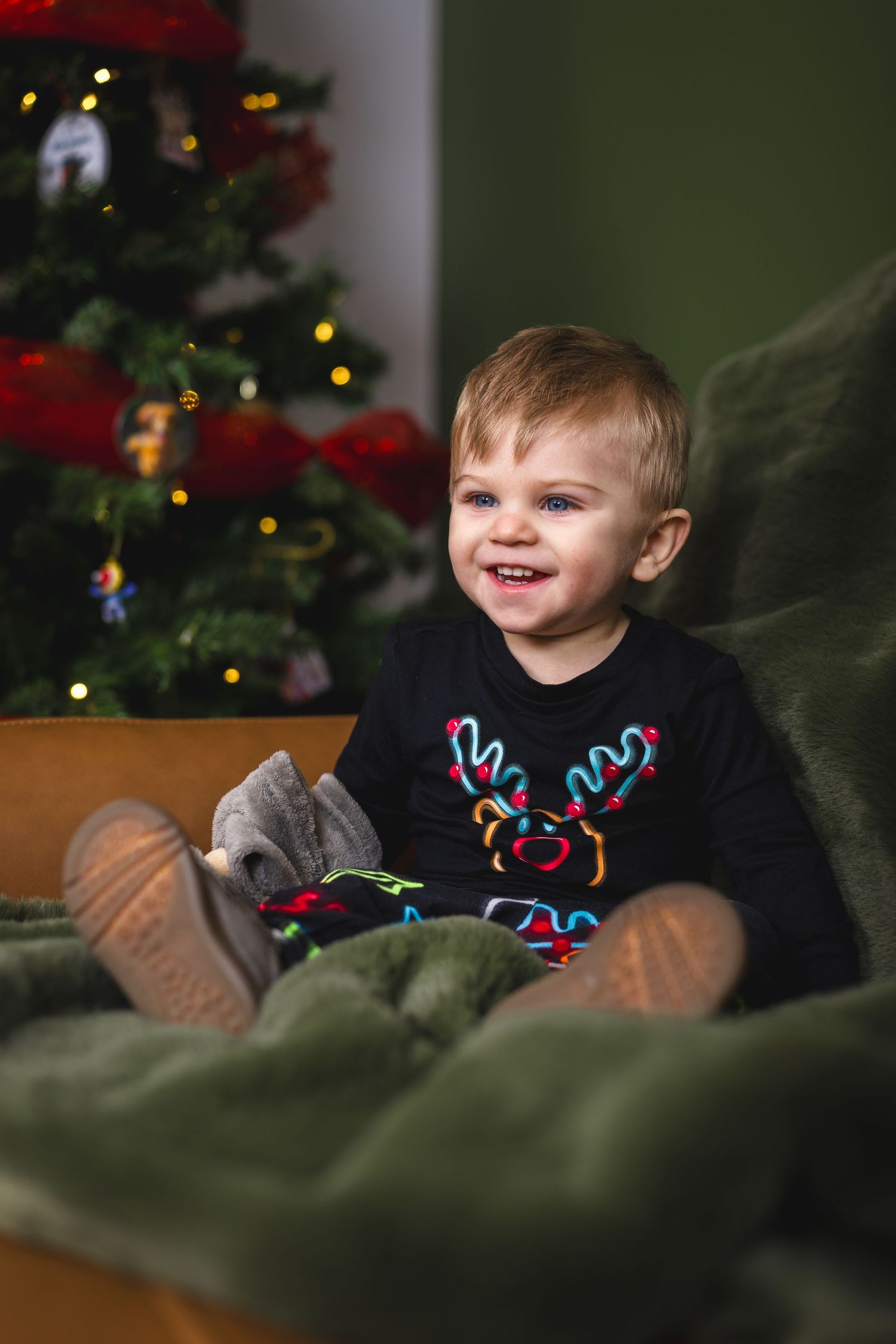 Child wearing holiday sweater sits on green couch with Christmas tree lights twinkling in background.