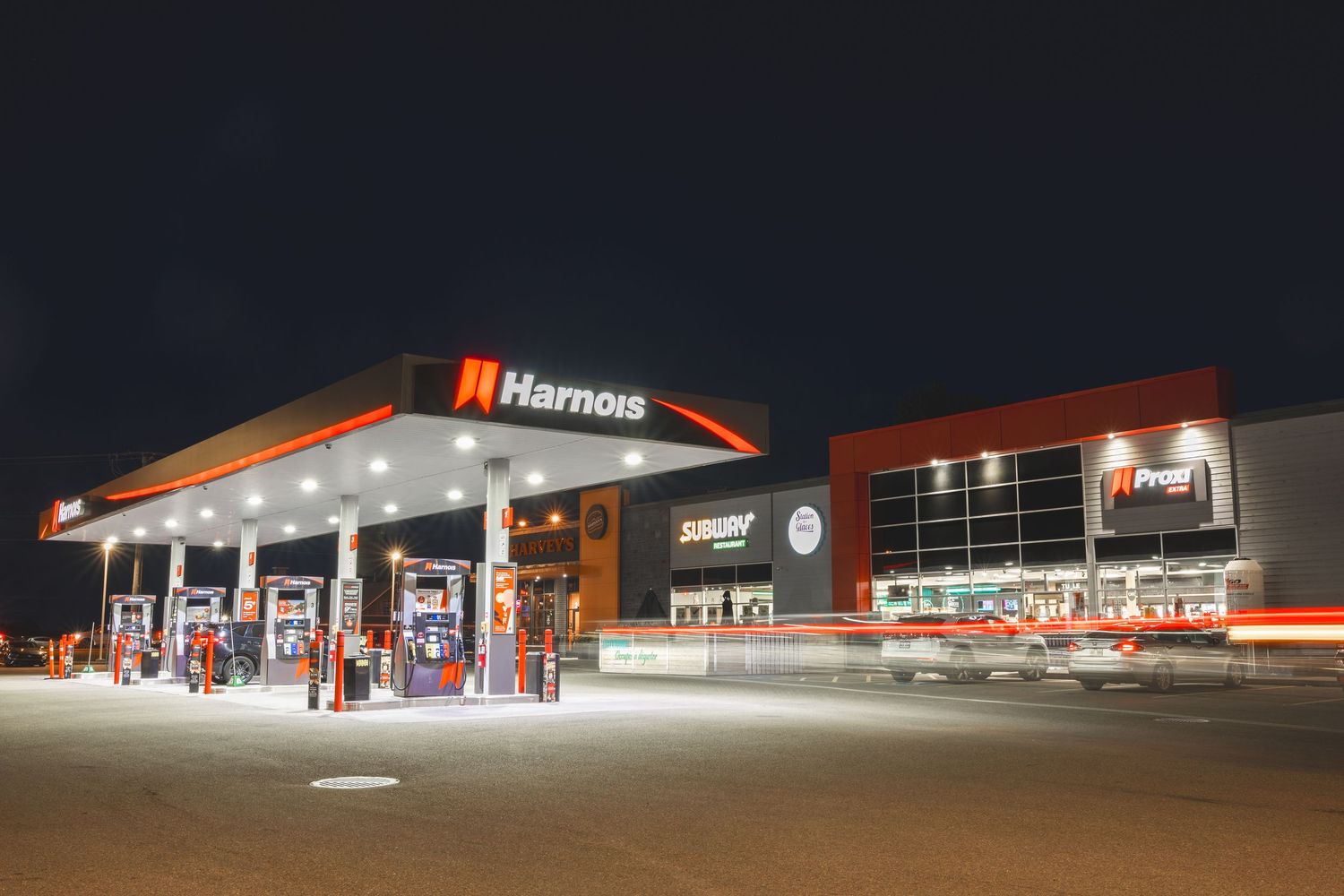 Illuminated Harmons grocery store exterior at night with bright red signage and multiple storefronts visible.