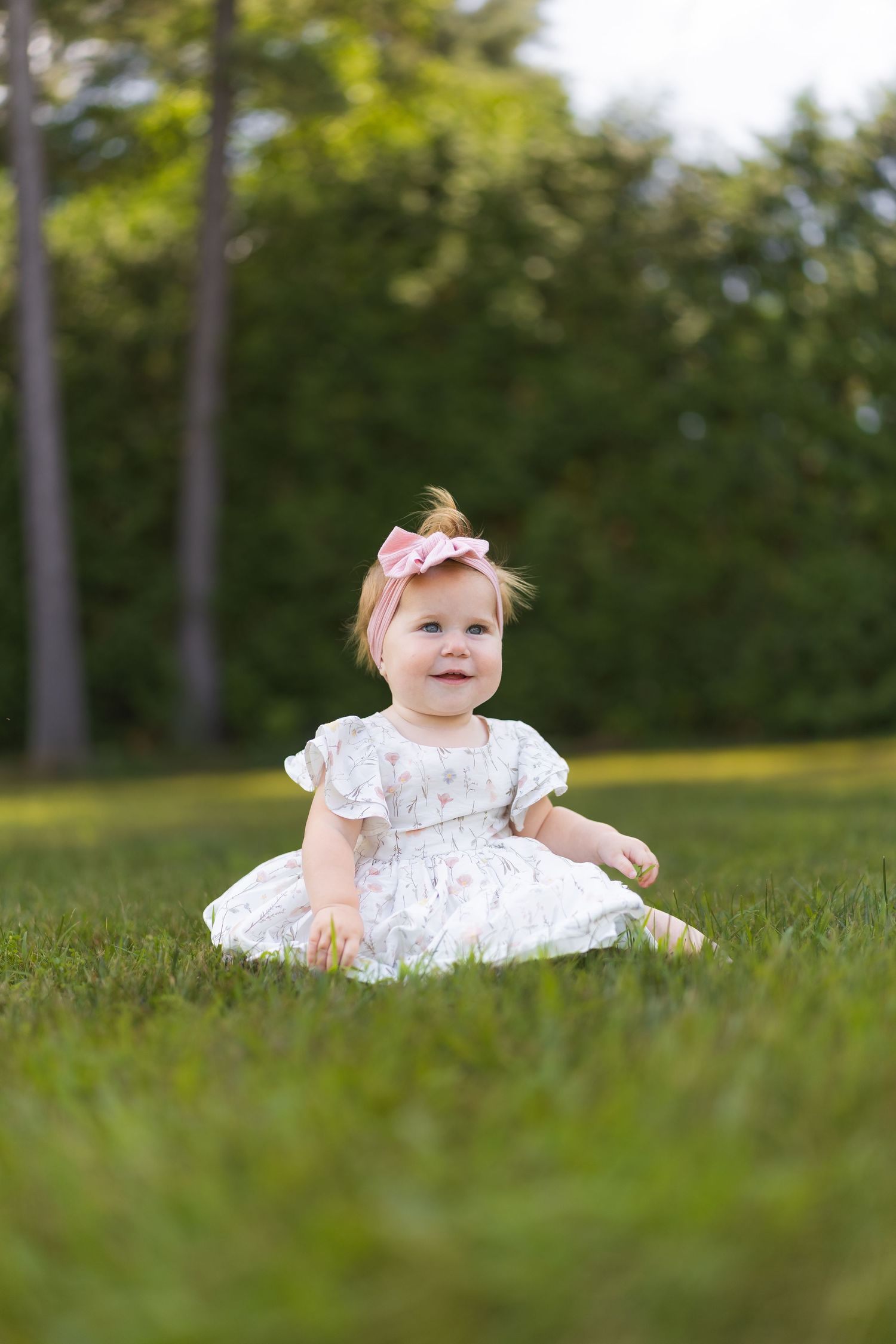 Smiling baby girl in white floral dress and pink bow headband sitting on green grass in a sunny outdoor park.