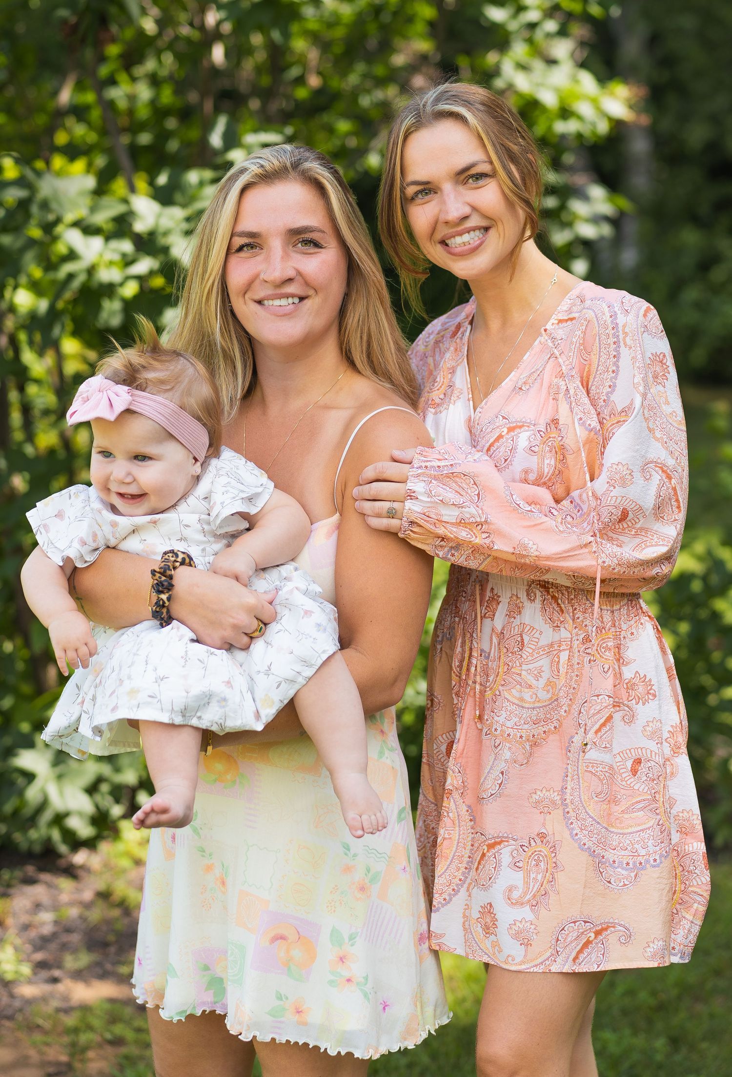 Two smiling women pose outdoors, one holding a baby girl in a white dress with a pink headband.