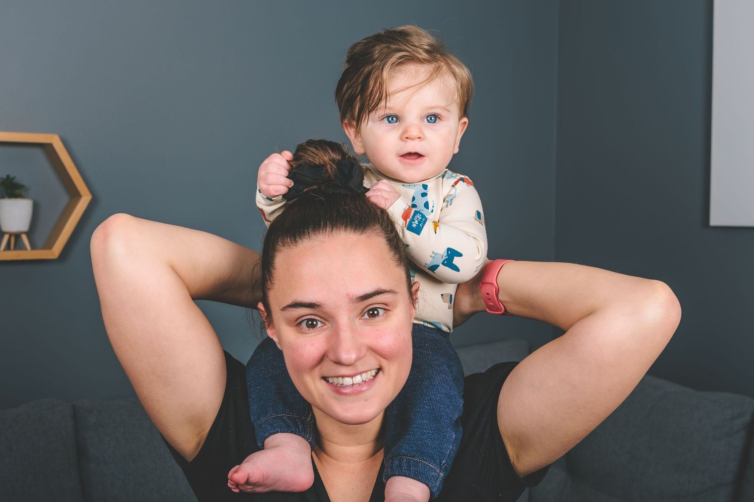 A playful moment captured as a baby sits on a parent's shoulders against a blue wall.