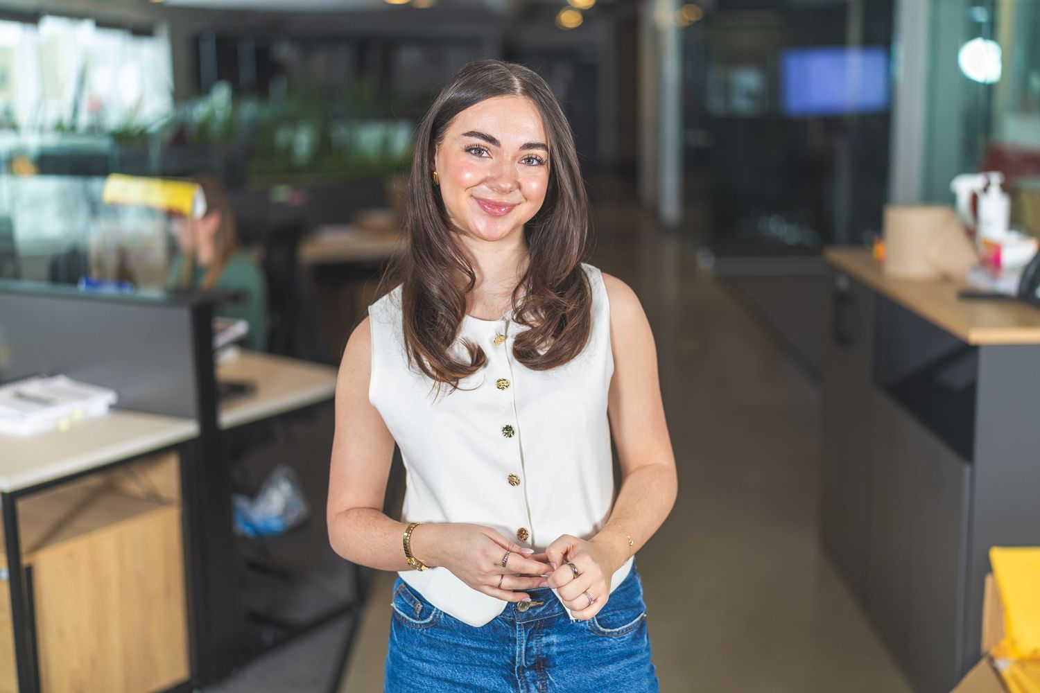 Professional in white sleeveless top and jeans stands in modern office space.
