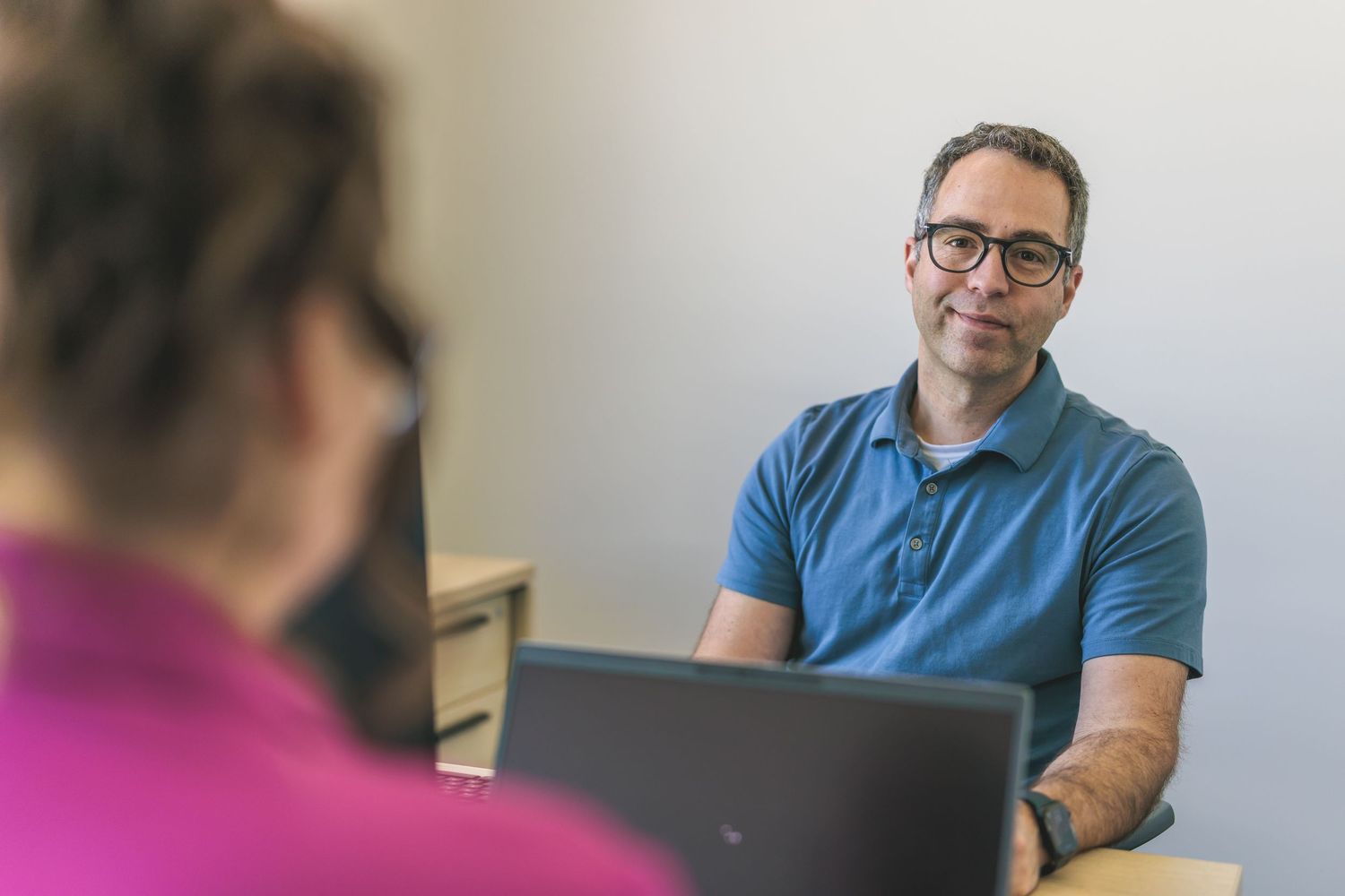 Office meeting scene with employee in blue polo shirt working on laptop.