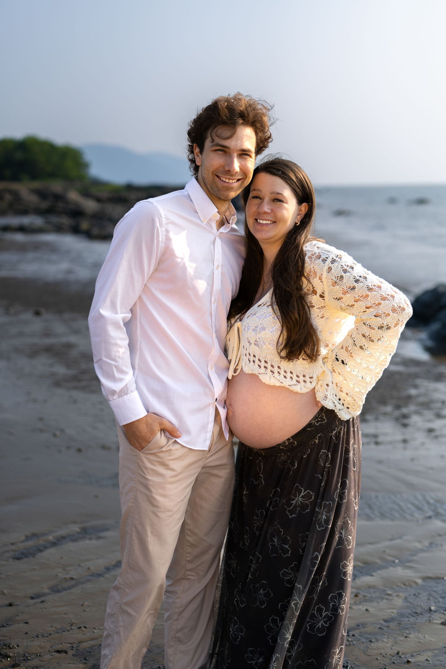 Smiling couple poses on beach, pregnant woman in crochet top and floral skirt with partner in white shirt.