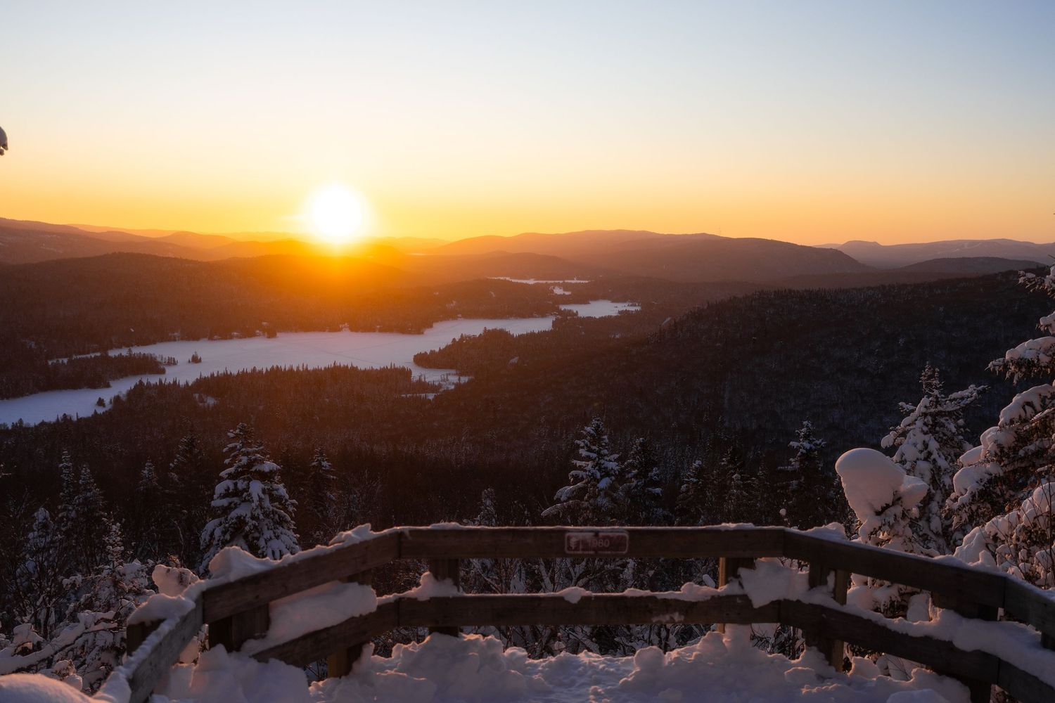 Panoramic winter sunset view from observation deck over snow-covered mountains and frozen lake.