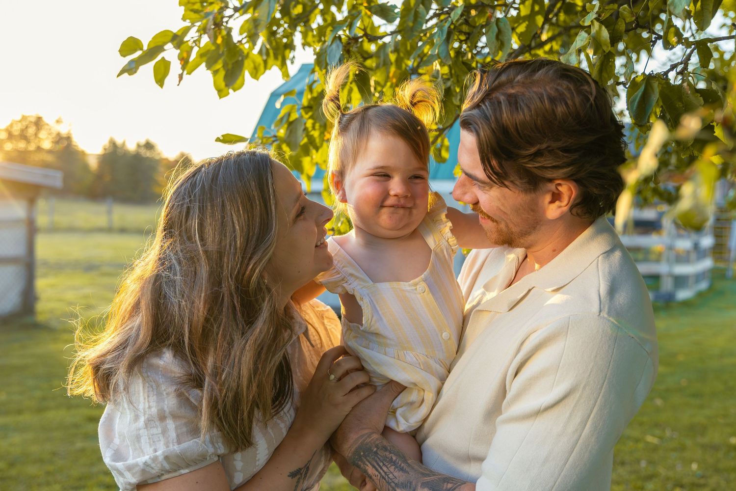 Family embraces while standing in a grassy field with trees and golden sunlight in the background.