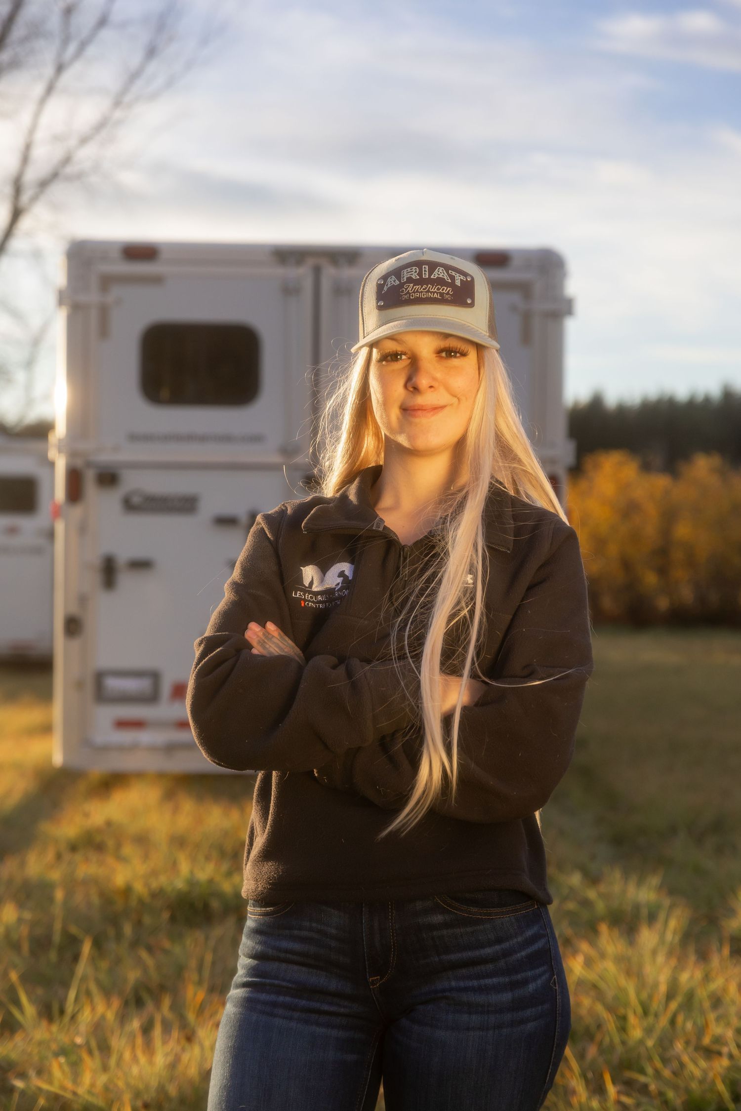 Person in black clothing and baseball cap poses near RV trailer in golden sunset field.