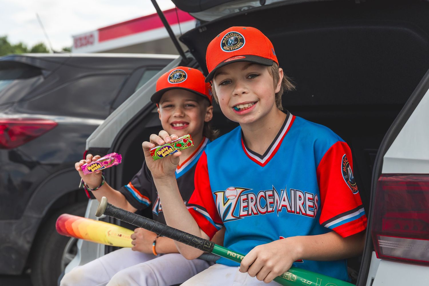 Two youth baseball players in red and blue uniforms enjoy snacks while sitting in the back of a vehicle.