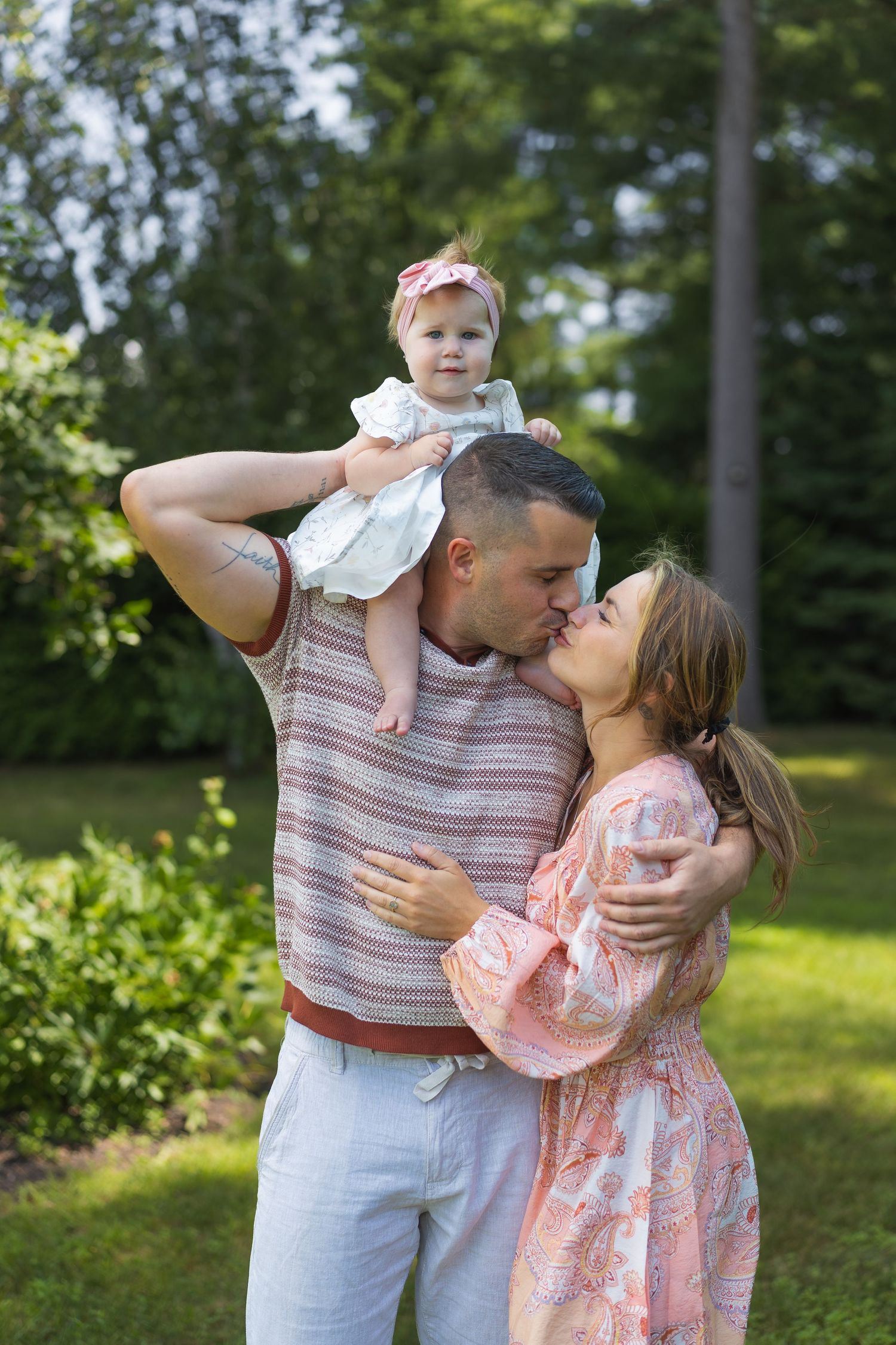Happy young family with baby girl sharing a kiss outdoors in a lush green garden on a sunny day.