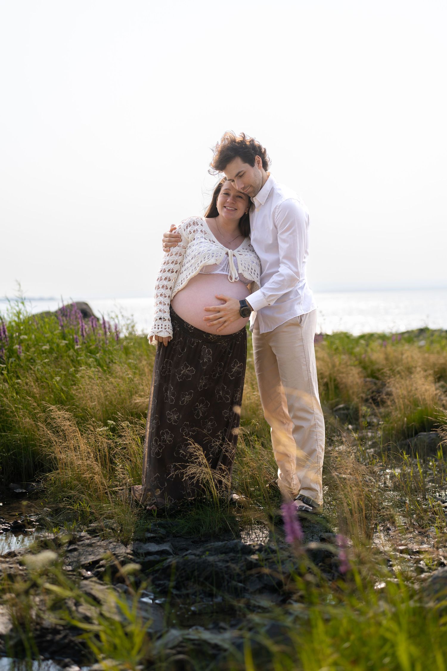 Couple embracing outdoors near water, pregnant woman showing baby bump, standing in lush green grass on a sunny day.