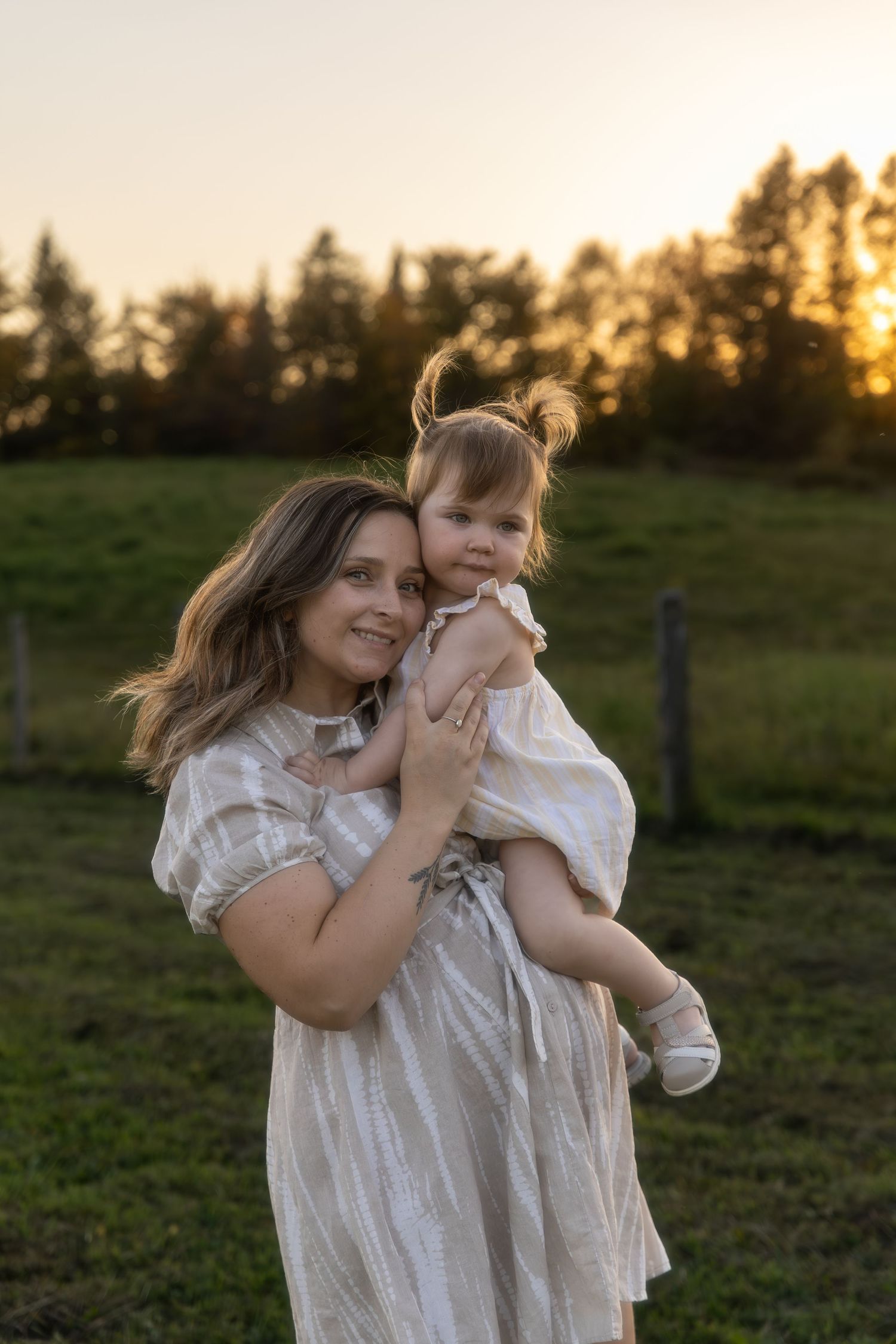Mother and child share playful moments in white dresses against a sunset backdrop in a green field.