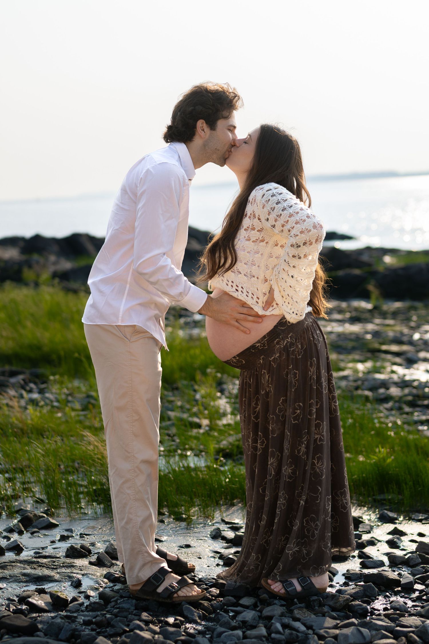 Couple kissing on rocky beach shore, pregnant woman in crochet sweater and brown skirt, man in white shirt.