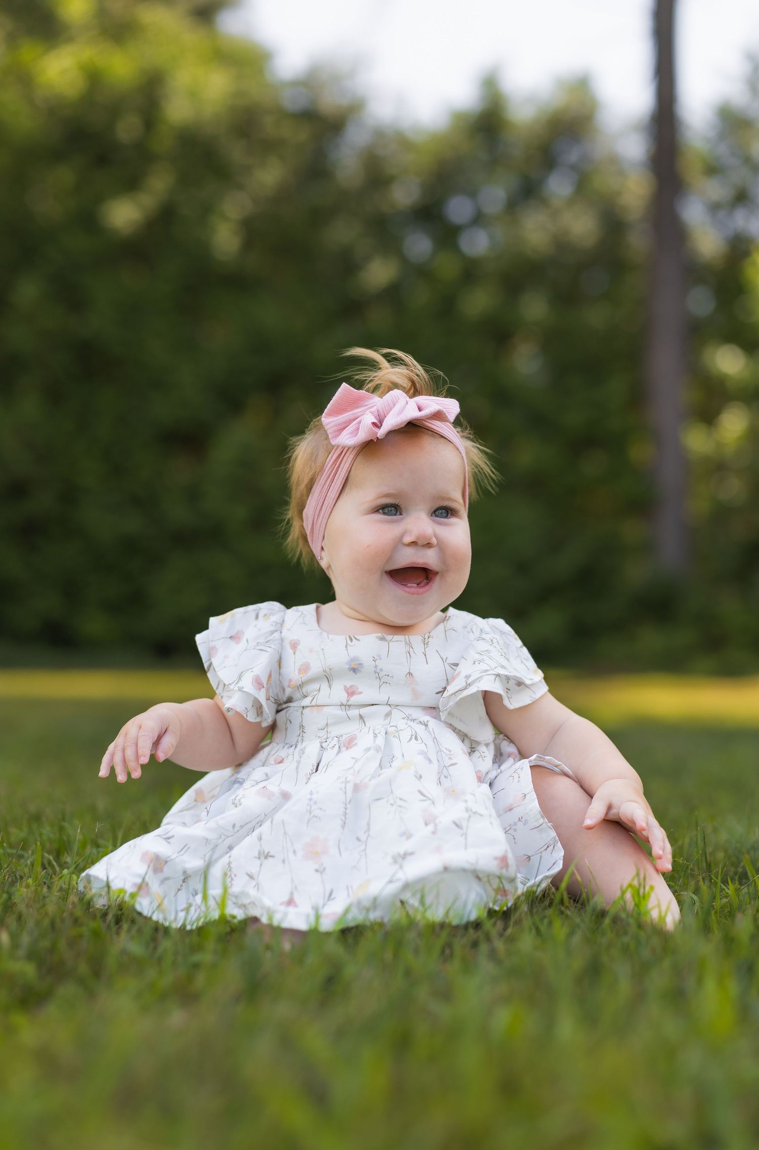 Smiling baby girl in floral dress and pink bow headband sitting on green grass outdoors.