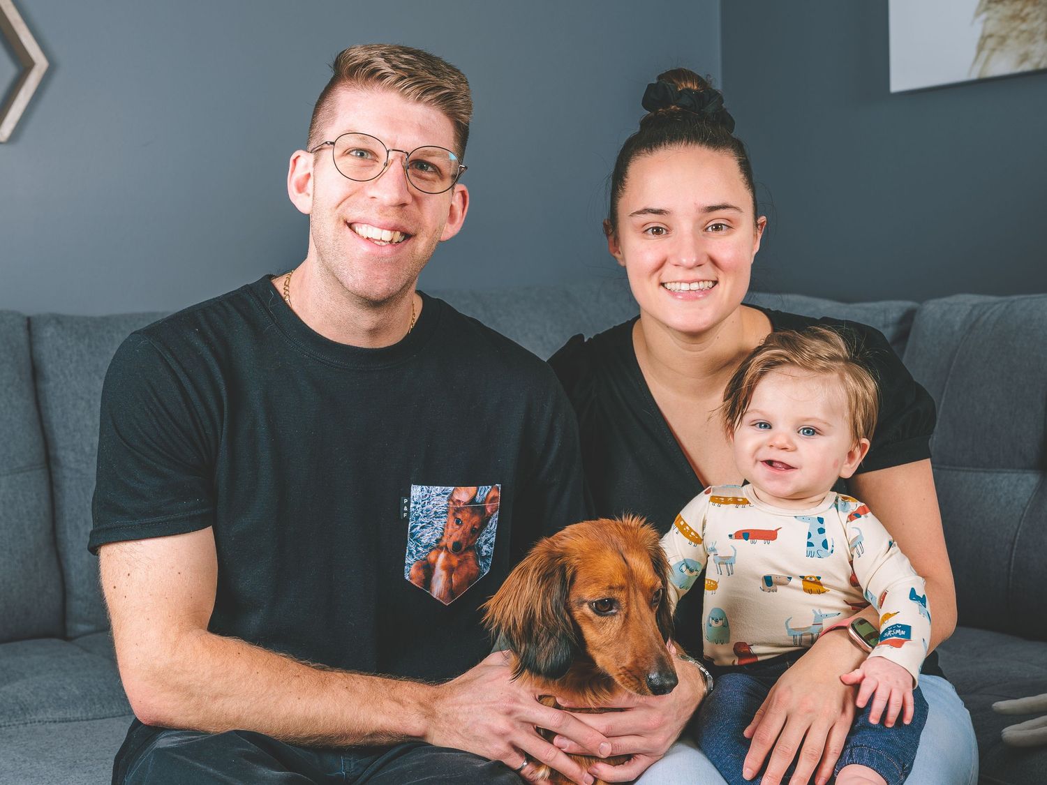 A happy family sits together on their couch with their brown dachshund pet dog.