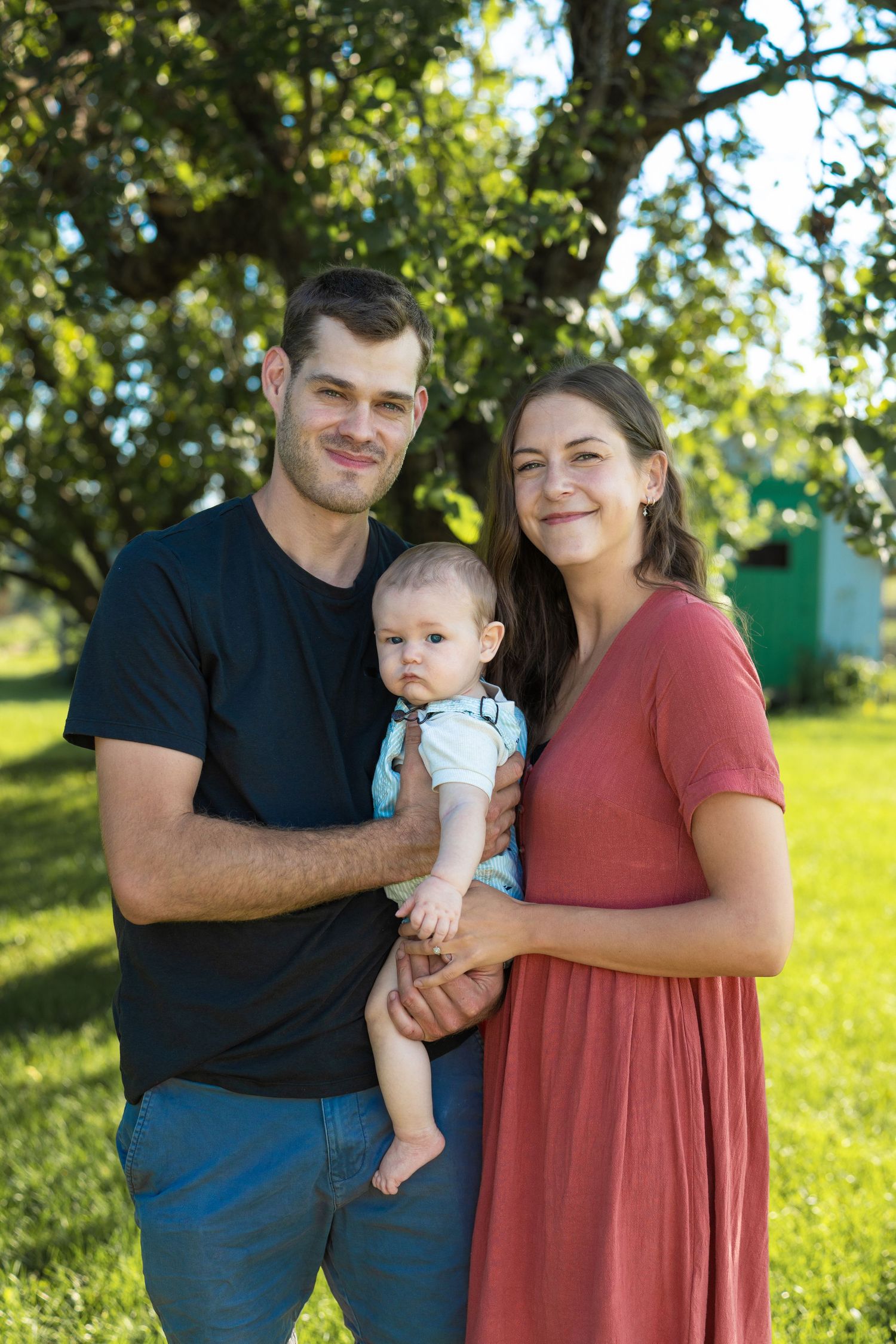 Young family with baby boy posing outdoors in green park, smiling couple holding infant child in summer garden.