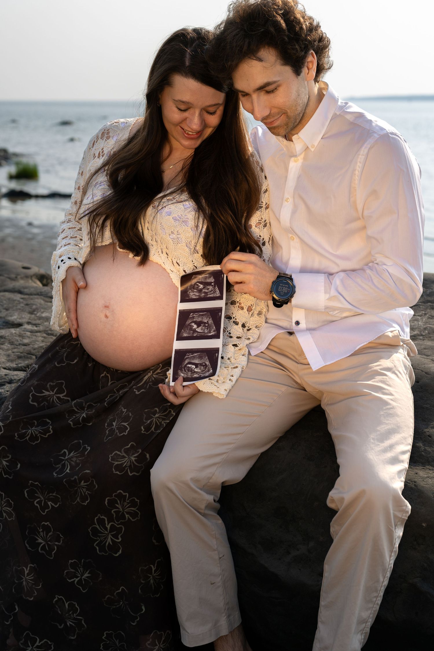 Pregnant couple sitting on rocks by the ocean, smiling while holding ultrasound photos together.