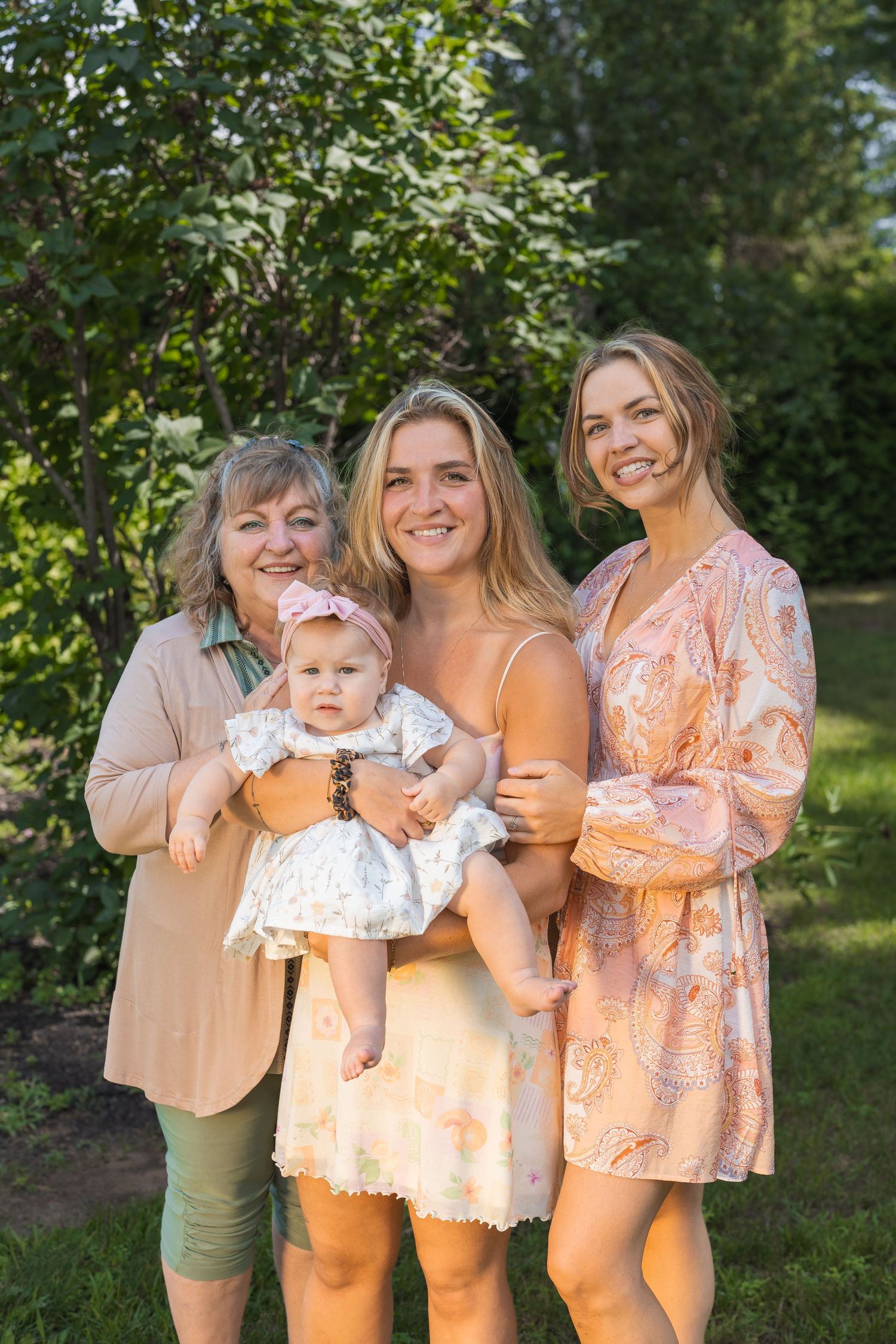 Three generations of women pose outdoors, with the middle woman holding a baby girl in a white dress.