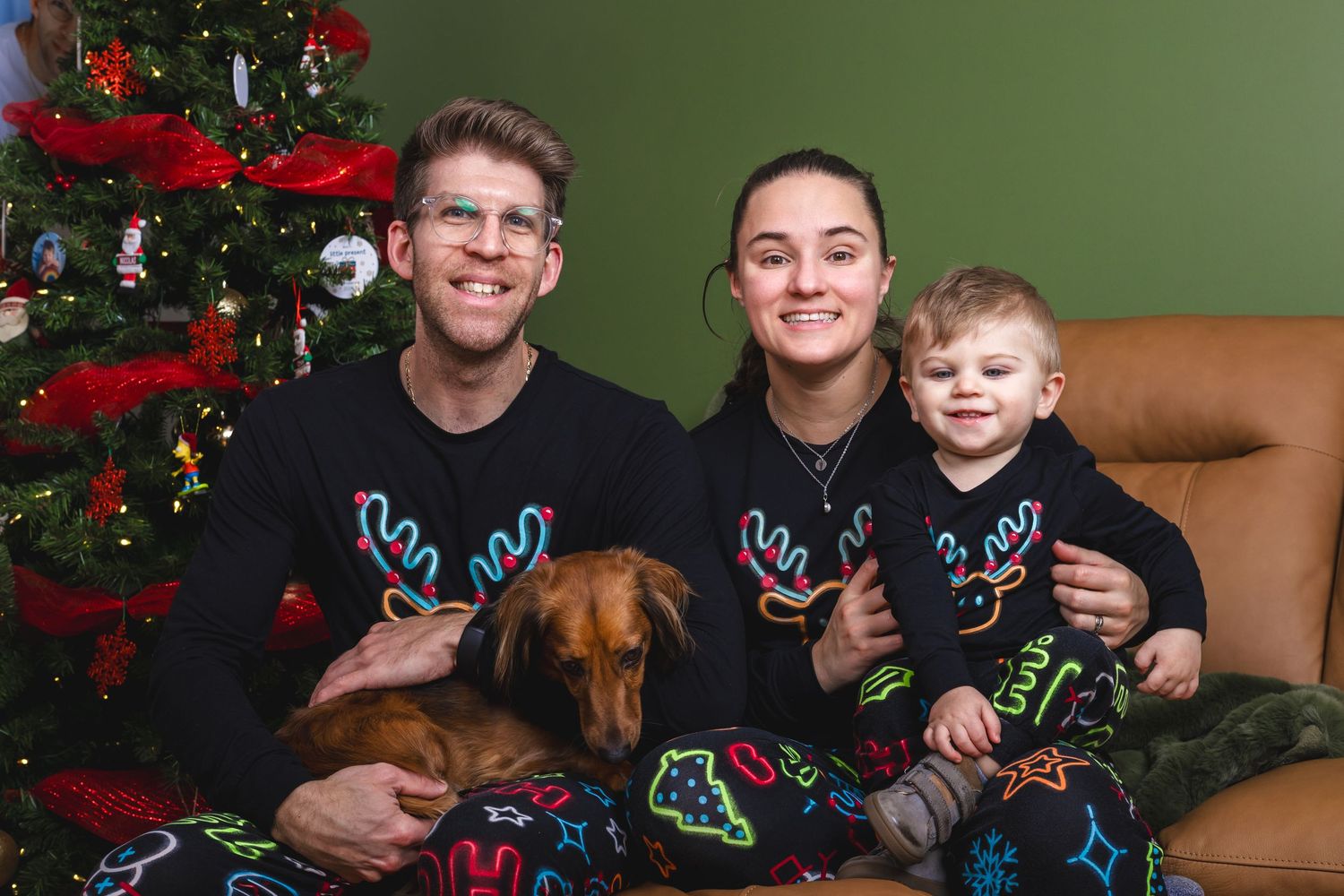 Family wearing matching holiday sweaters pose together on brown couch with Christmas tree.