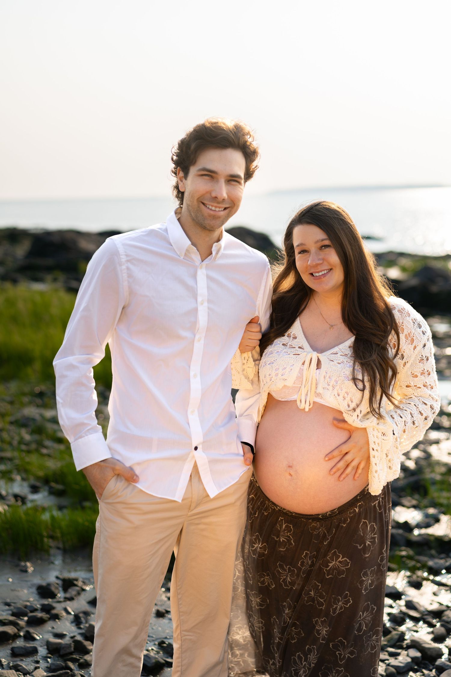 Smiling couple posing on rocky beach, pregnant woman in lace top holding baby bump, man in white shirt beside her.