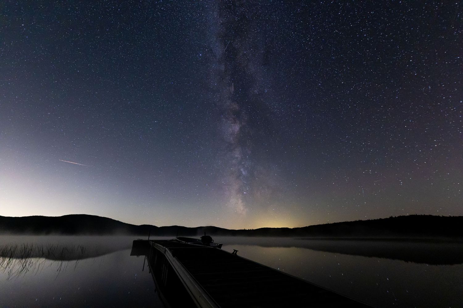 Milky Way galaxy stretching across night sky over a dark lake with wooden pier.