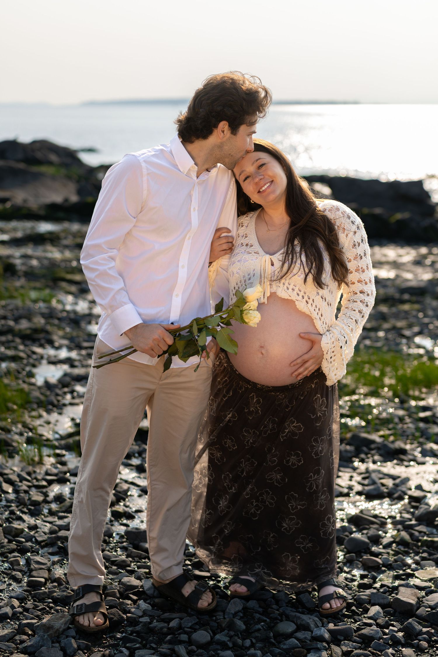A couple shares a tender moment on a rocky beach, with the pregnant woman cradling her belly and holding white roses.