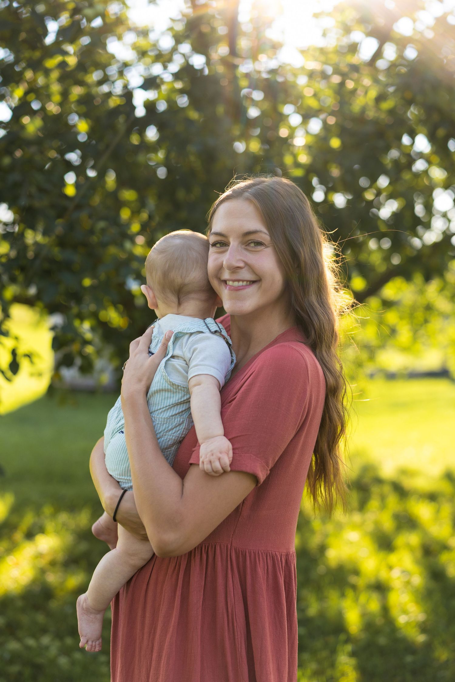 Young smiling mother in red dress holding baby outdoors in sunlit green park setting.